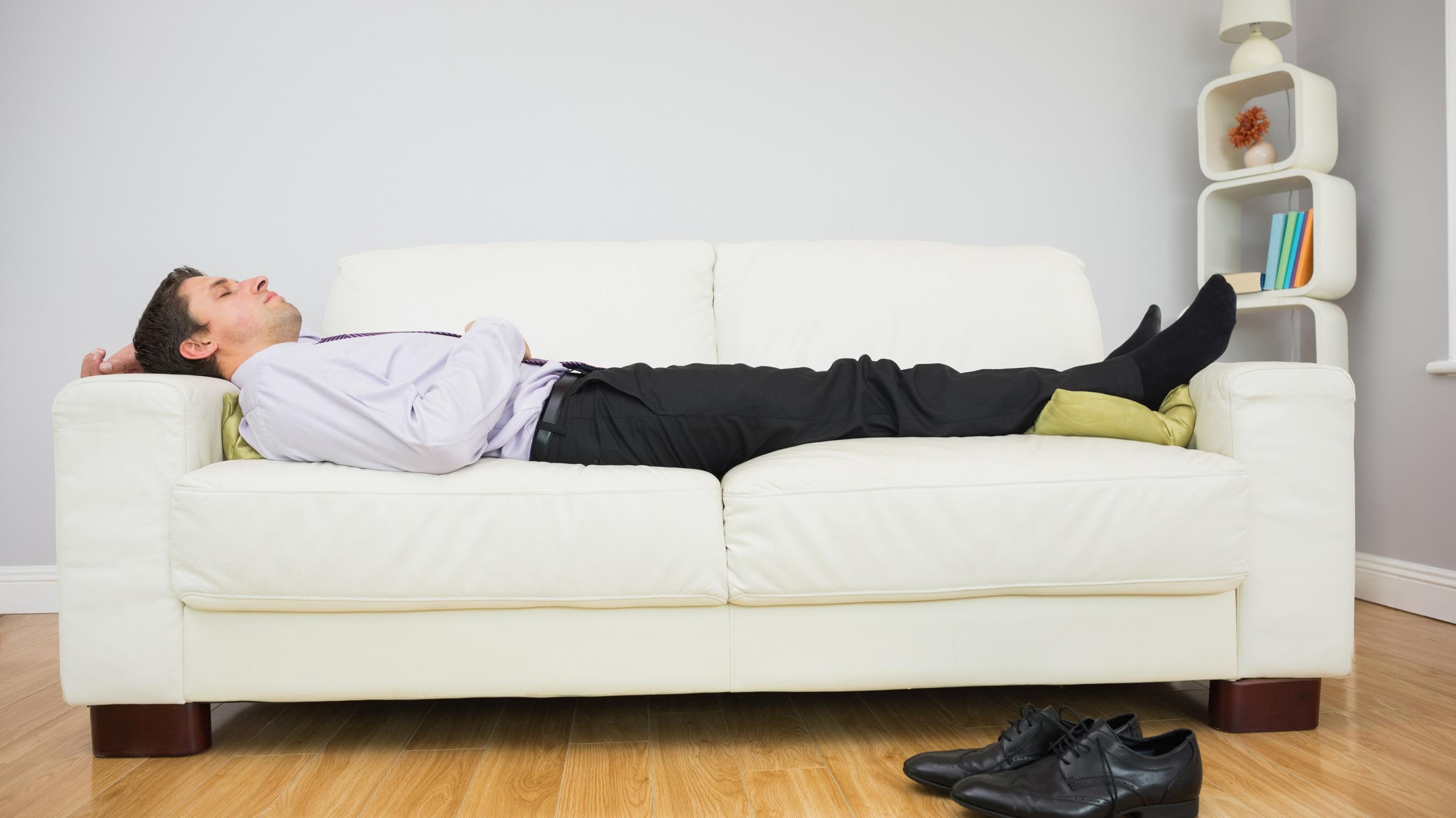 Ein Mann liegt ausgestreckt auf einem Sofa und schläft. Er hat noch Anzughose, Schlips und Hemd an, seine schwarzen Schuhe liegen auf dem Boden.
