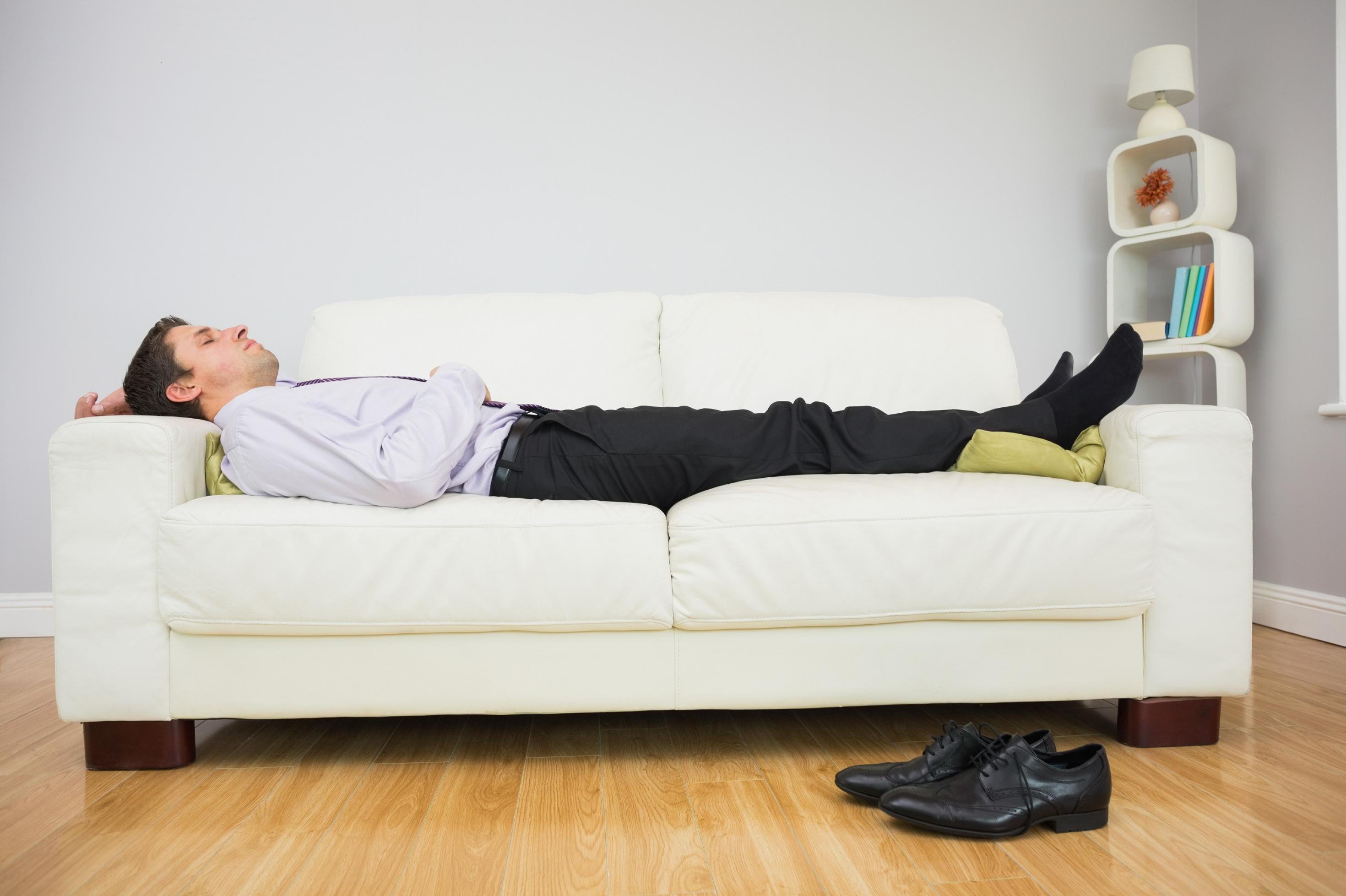 Ein Mann liegt ausgestreckt auf einem Sofa und schläft. Er hat noch Anzughose, Schlips und Hemd an, seine schwarzen Schuhe liegen auf dem Boden.