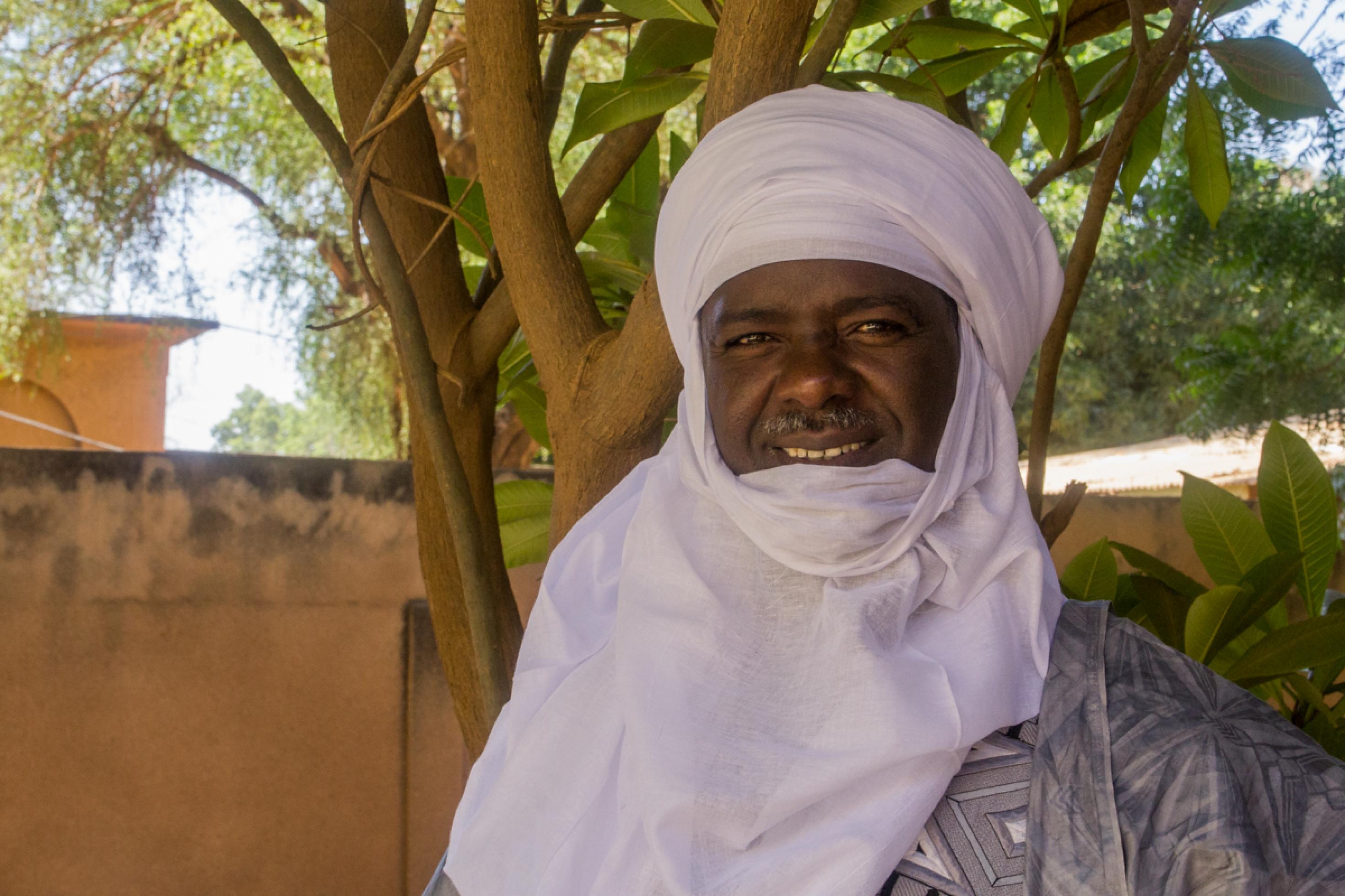 Portrait eines älteren Mannes mit einem weißen Tuch um Kopf und Hals. Er steht vor einem Baum