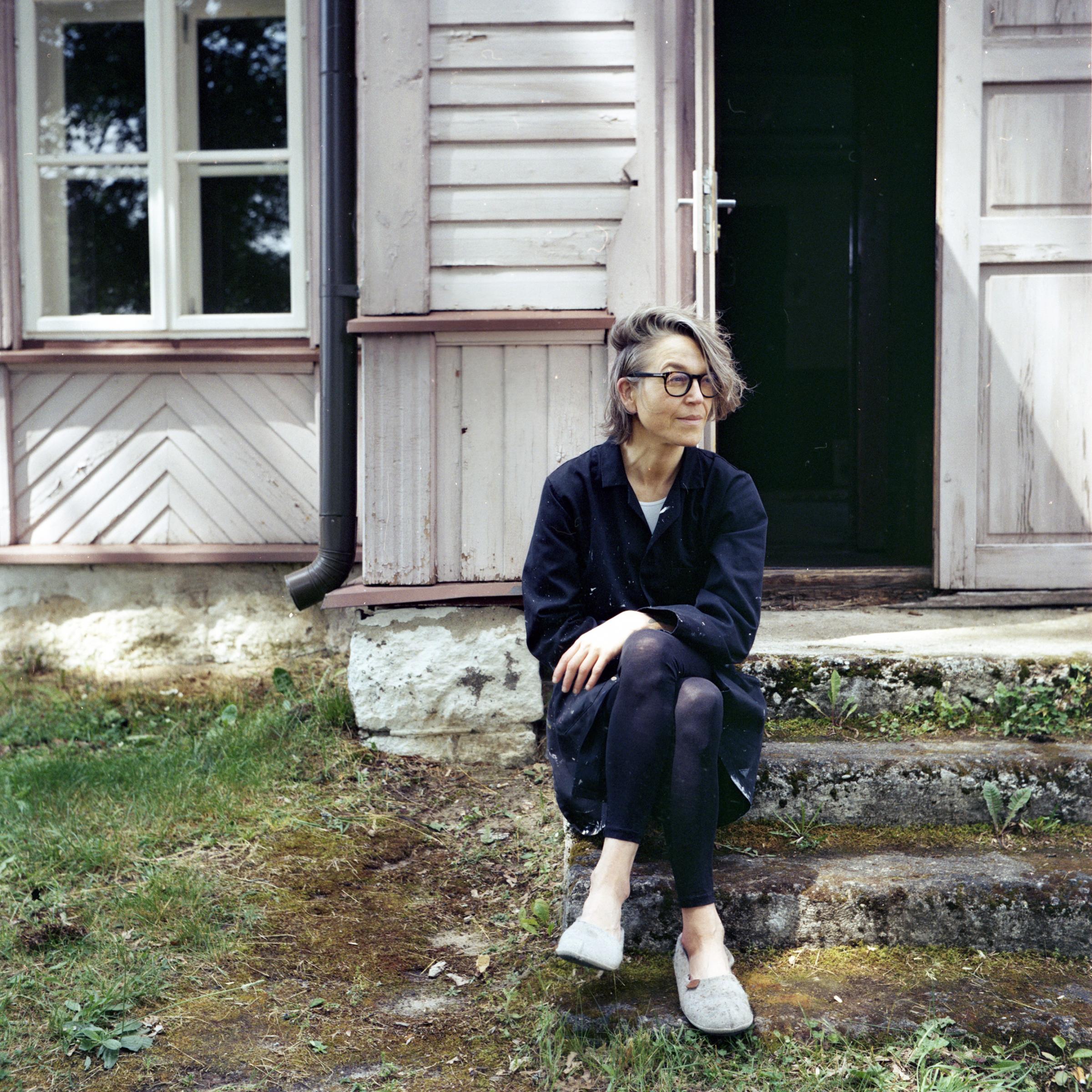 Frau mit grauem Haar sitzt lässig vor altem weißem Holzhaus auf den Stufen.