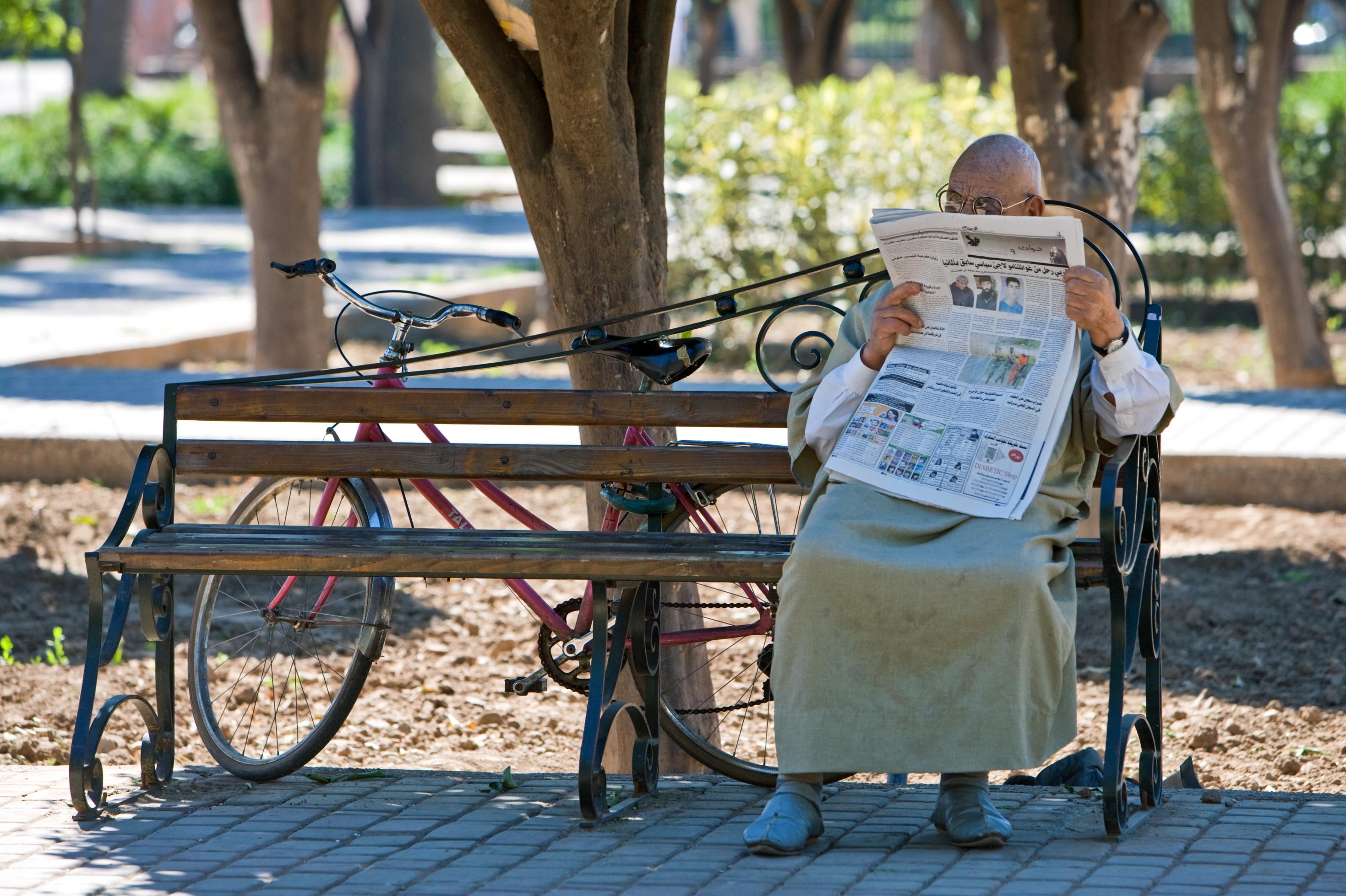 Ein älterer Mann sitzt auf einer Bank im Park und liest Zeitung. Im Hintergrund lehnt ein Fahrrad an einem Baum.