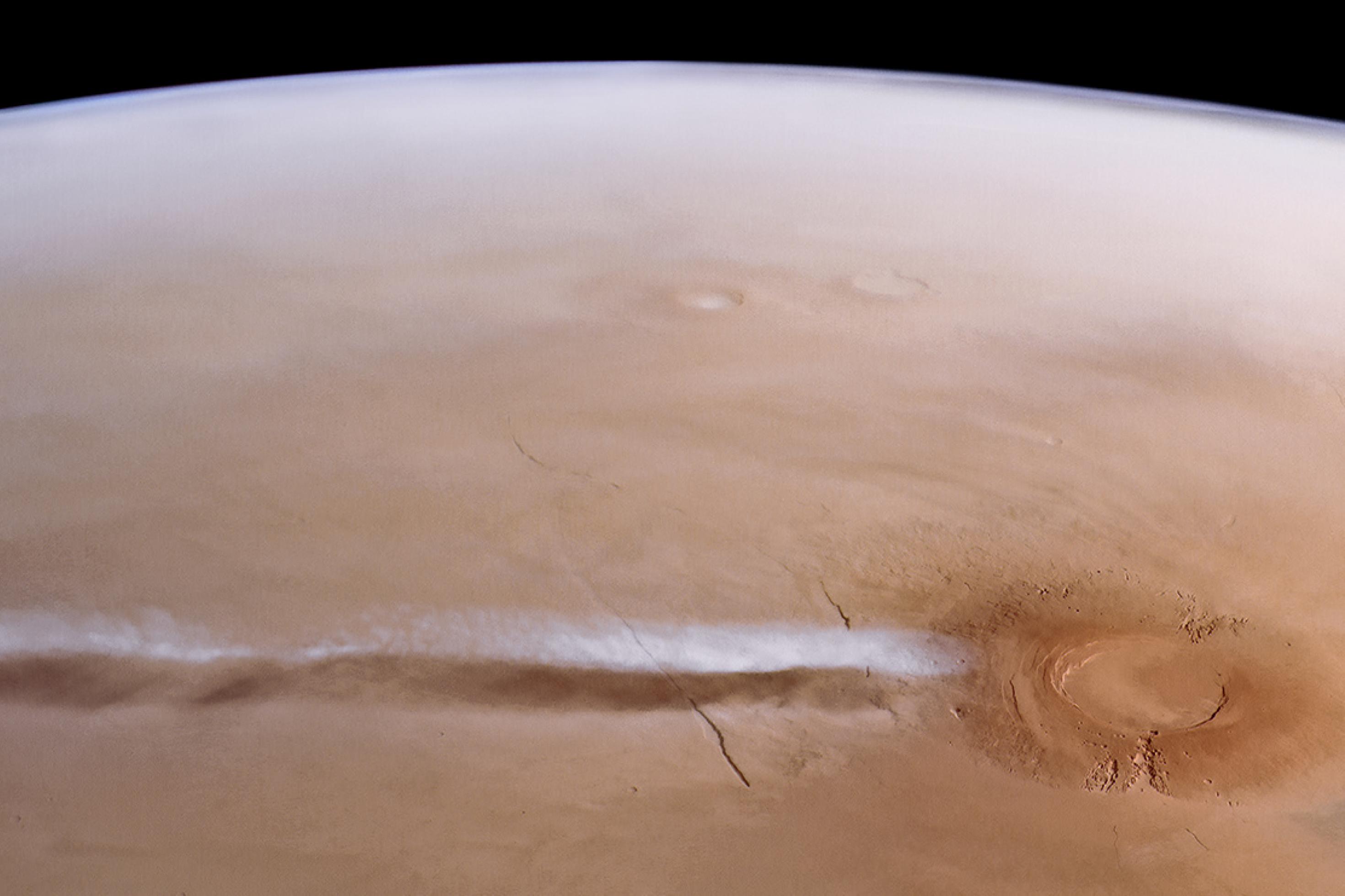 Der Marsvulkan aus dem Orbit: Ein runder Berg mit einer Einbuchtung, der Caldera. Darüber eine breite, langgestreckte Wolke.