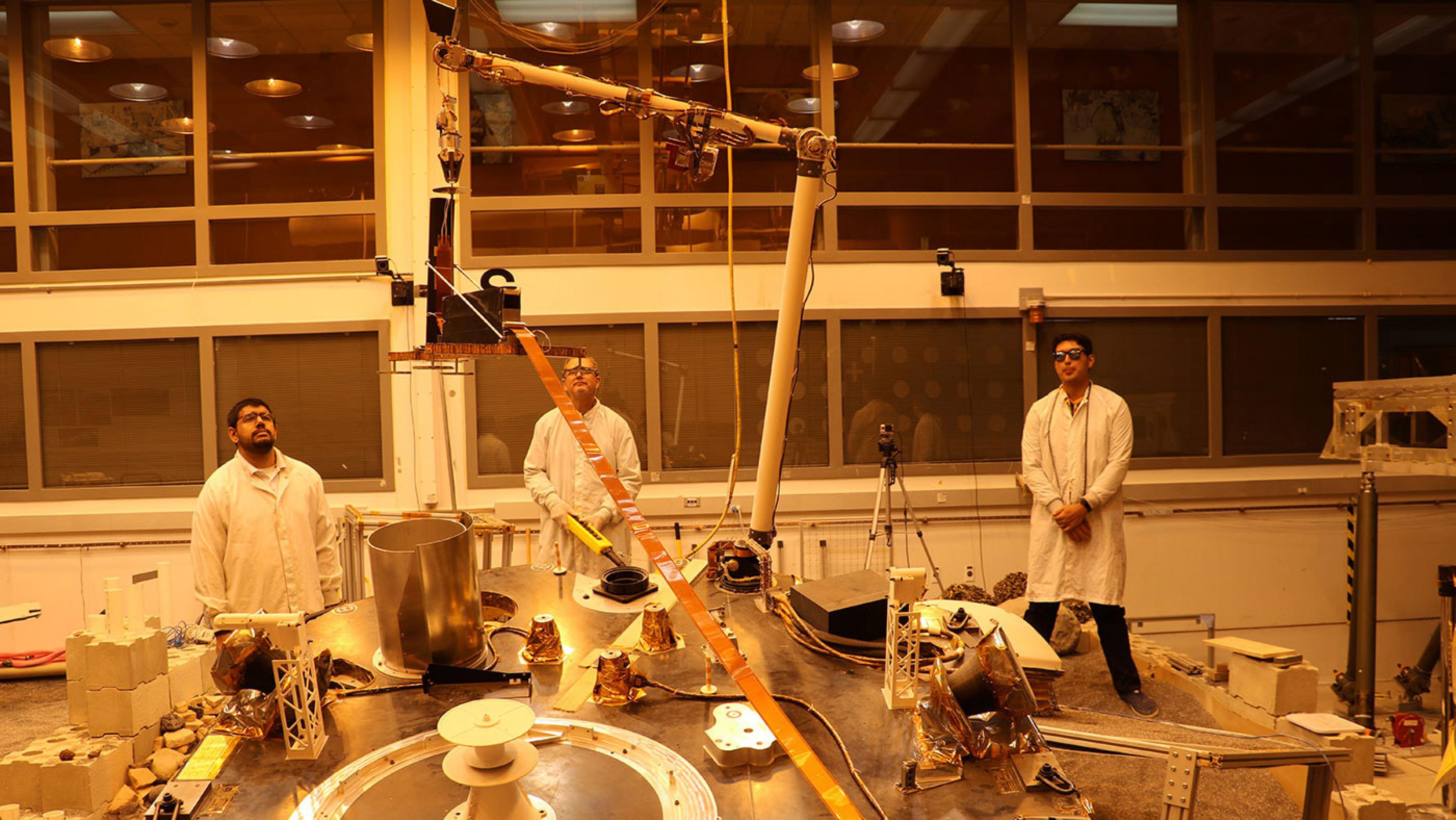 Ein Roboterarm in einem Labor auf der Erde hebt die Wärmesonde in die Höhe, während drei Forscher zusehen
