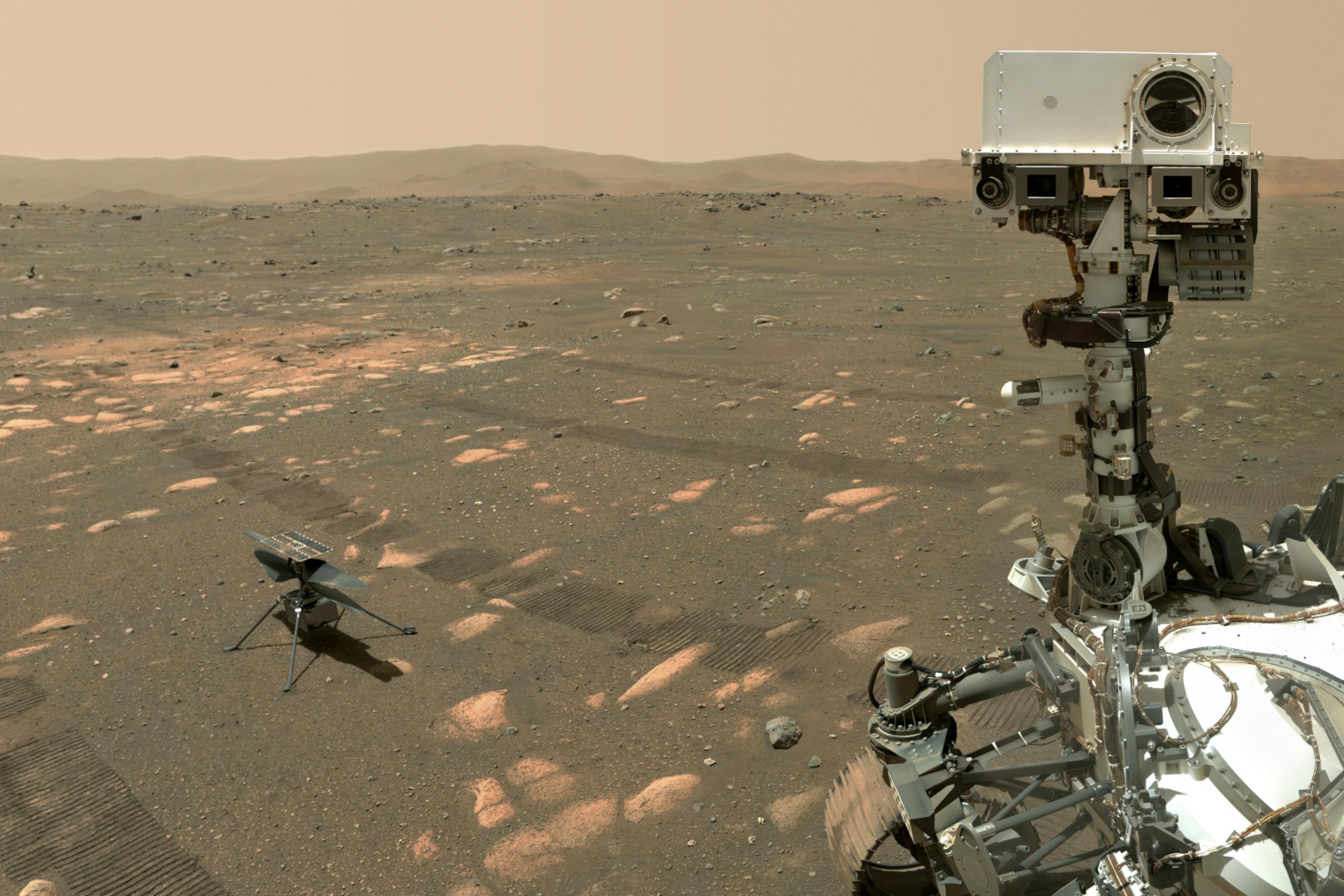 Perseverance steht in einer bräunlichen Ebene mit einigen flachen Steinen, am Horizont einige Hügel, der Rover „blickt“ zum Betrachter, neben ihm steht klein der Marshelikopter