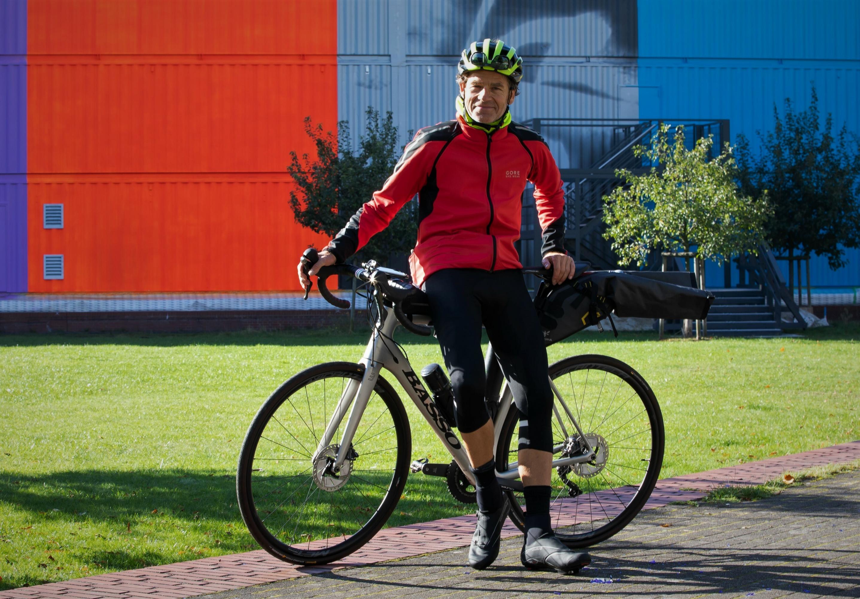 Martin C Roos mit seinem Rennrad vor einer sonnenüberfluteten Wiese, hinter der bunt gestrichene Metallwände aufragen