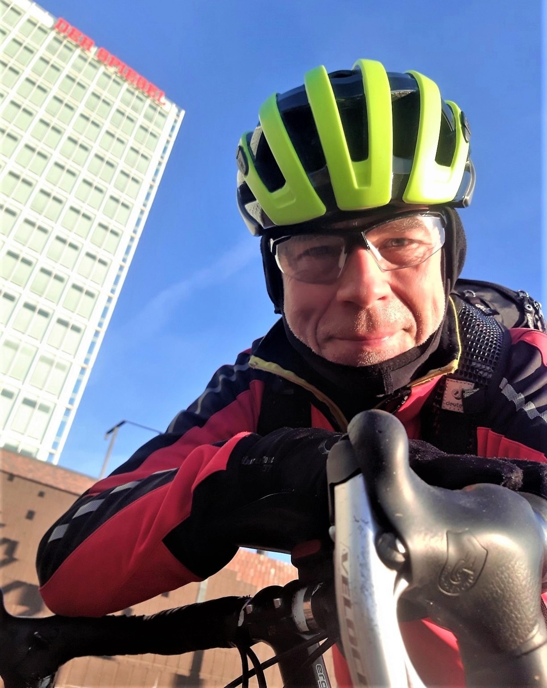 Der behelmte und dick eingemummelte Reporter lugt über den Lenker seines Rennrads; im Hintergrund ragt das vom roten Schriftzug „Der Spiegel“ gekrönte Hochhaus in einen stahlblauen Himmel
