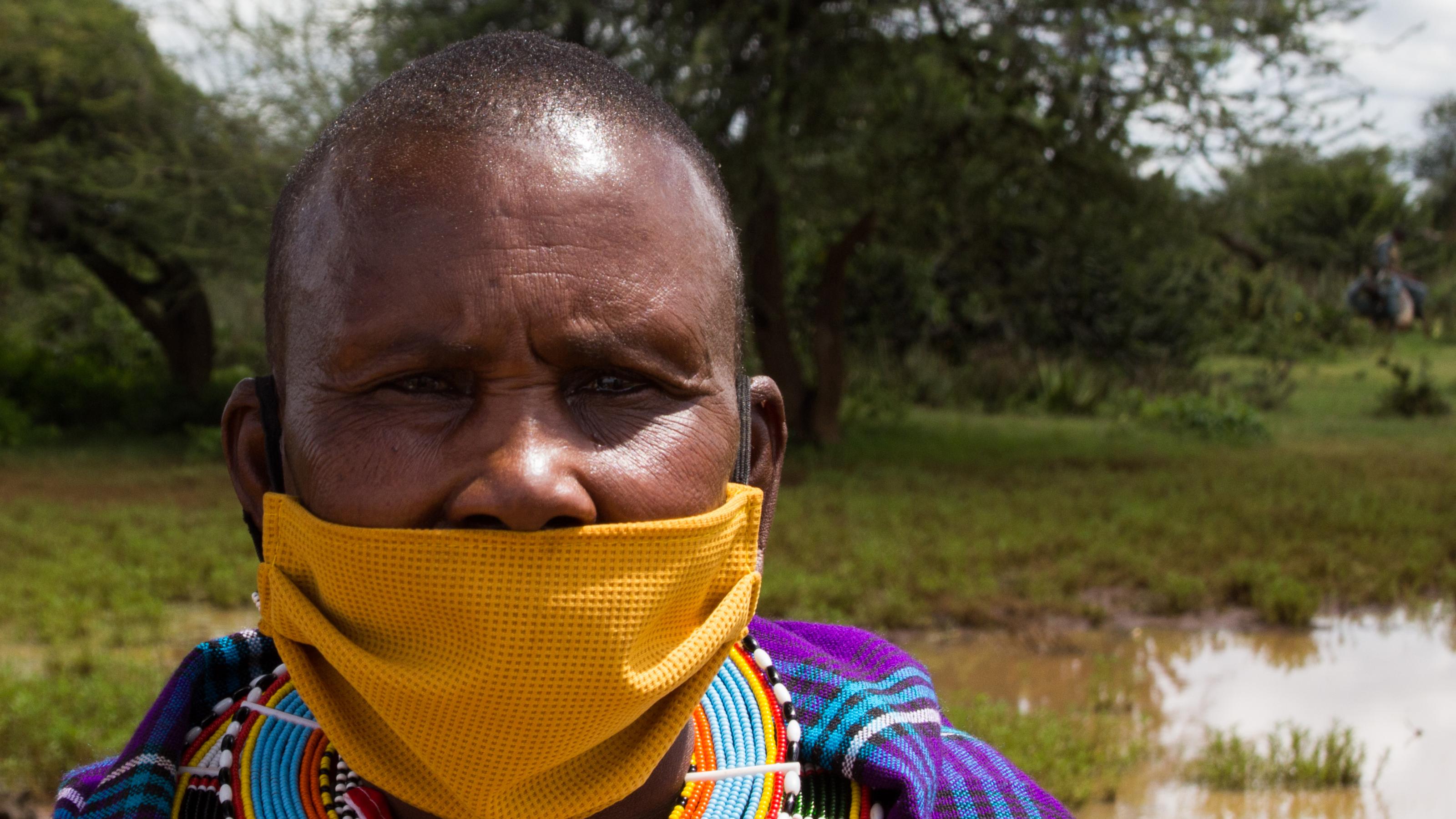 Zu sehen ist eine Massai-Frau in traditioneller Kleidung, d.h. in einem kräftig blauen Kleid mit reichlich Perlenschmuck um den Hals. Sie trägt einen gelben Mundschutz, wobei sie die Nase nicht bedeckt hat.