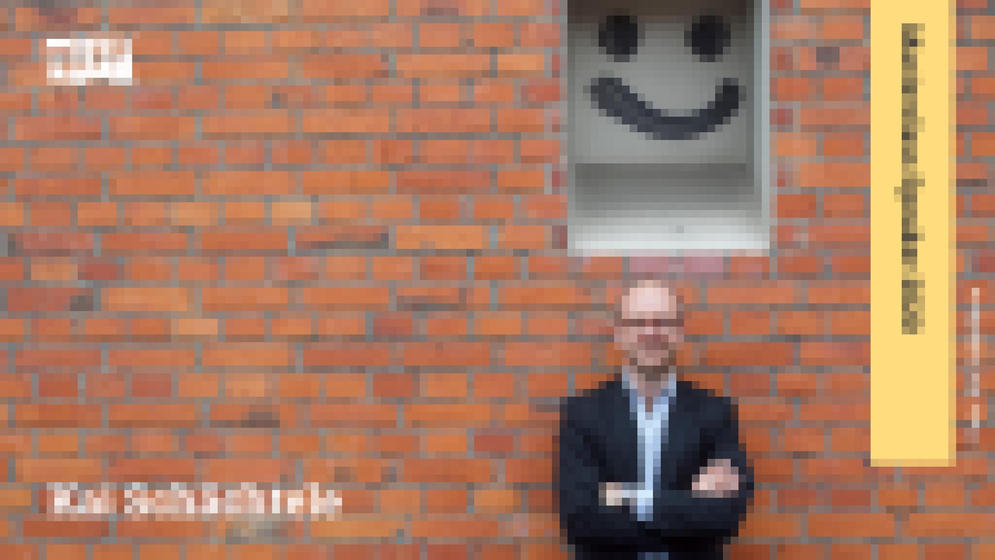 Ein Mann mit Brille steht vor einer roten Ziegelmauer. Über ihm ist die Mauer weiß und trägt ein mit Sprühfarbe aufgemalten Smiley.