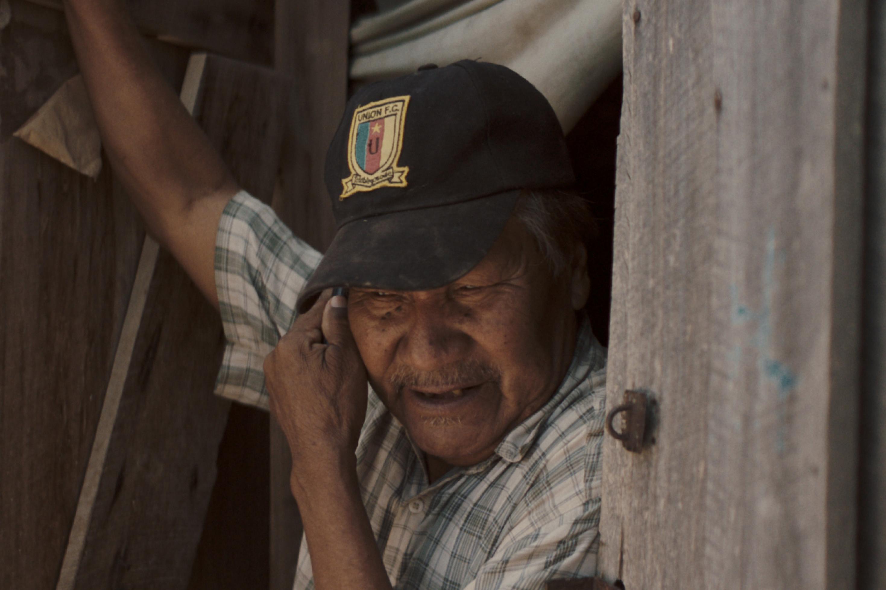 Ein etwa 70jähriger indigener Mann steht im Türrahmen seines Holzhauses der staubigen Siedlung „Campo Loro“, Paraguay.  Er blickt nachdenklich und hat eine Baseball-Kappe auf.