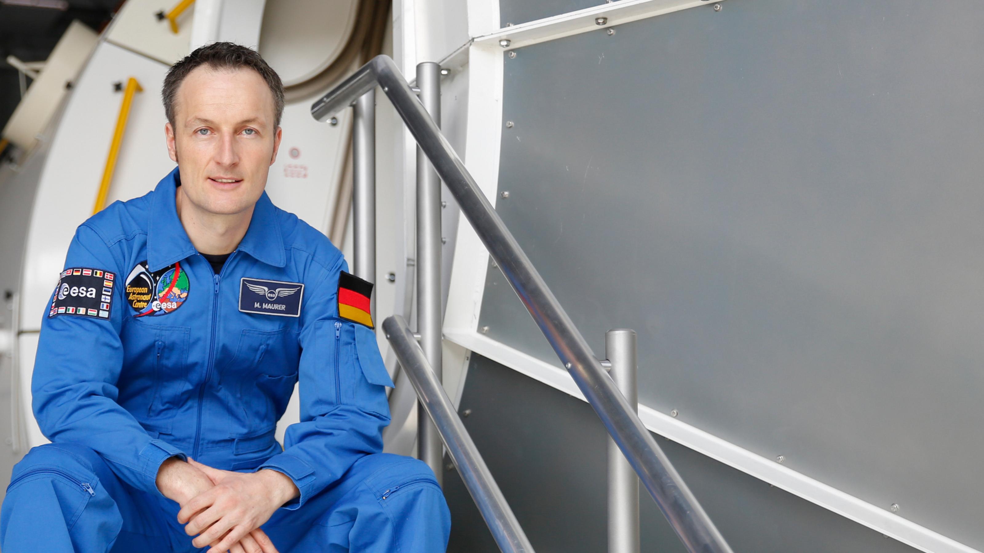 Der Astronaut Matthias Maurer sitzt in blauem Anzug auf einer Treppe vor dem Columbus-Modul