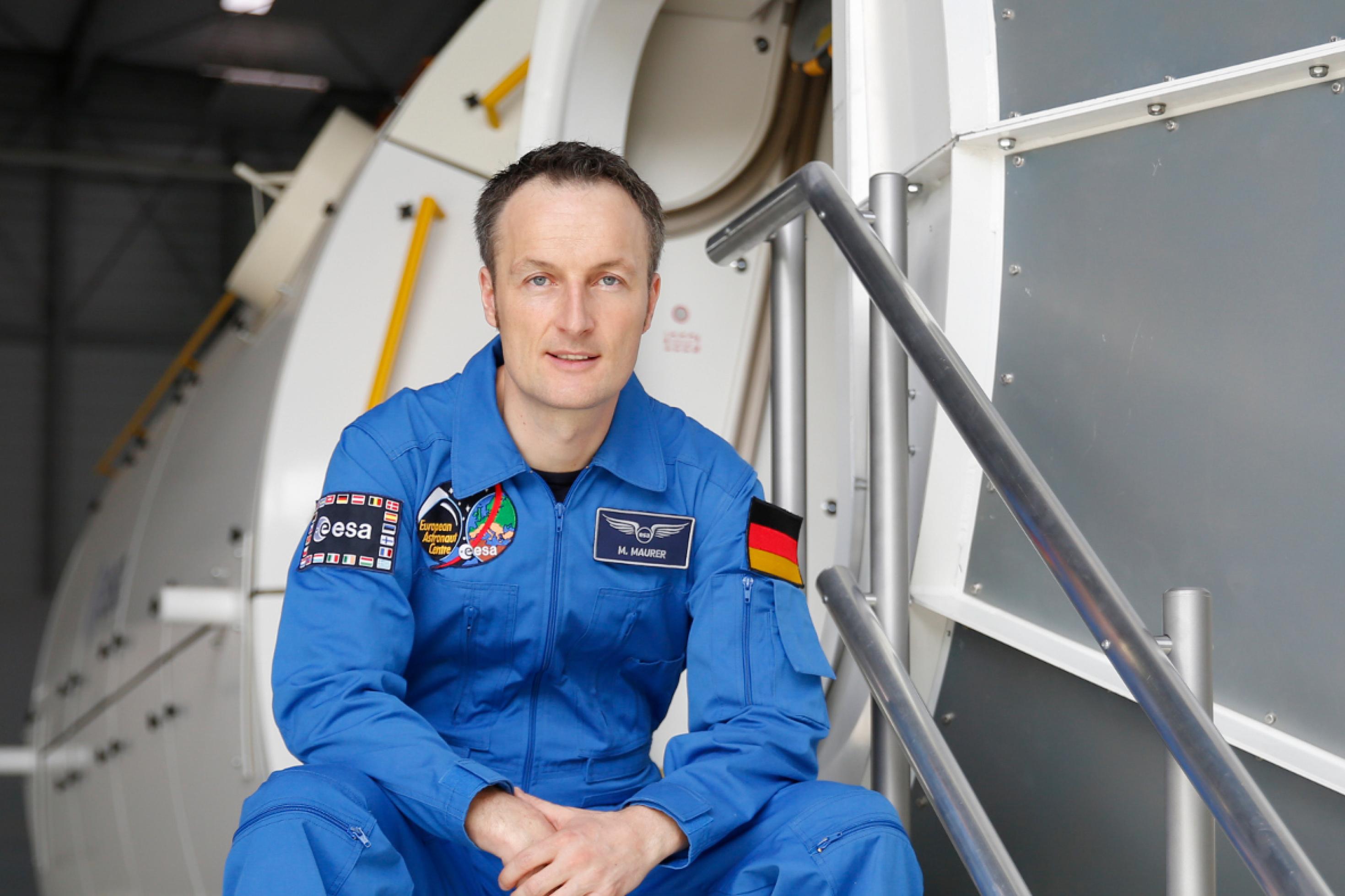 Der Astronaut Matthias Maurer sitzt in blauem Anzug auf einer Treppe vor dem Columbus-Modul
