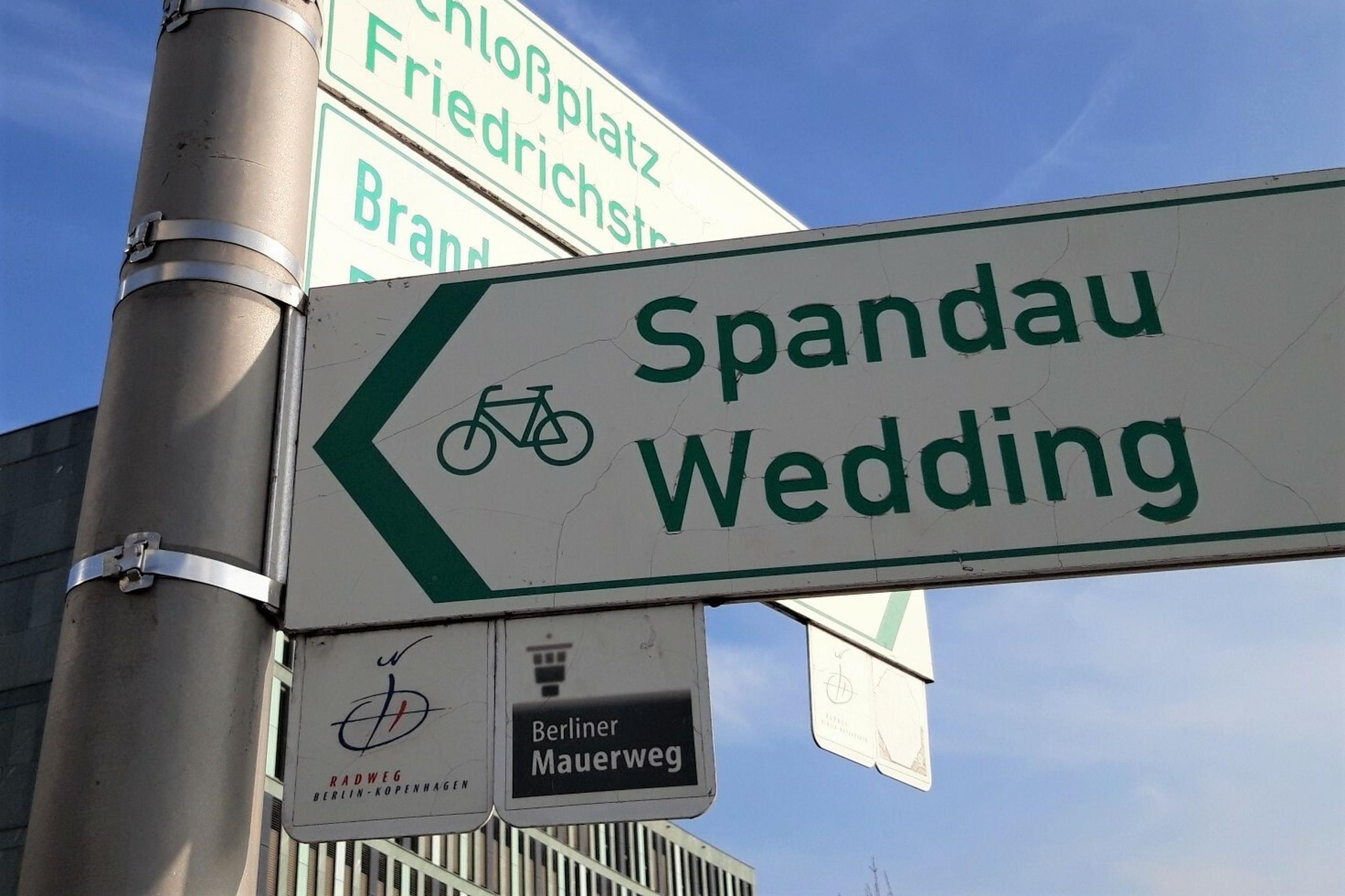 Unter dem großen Radrouten-Hinweisschild mit der Aufschrift „Spandau, Wedding“ steht klein und weiß, auf braunem Grund: „Berliner Mauerweg“.