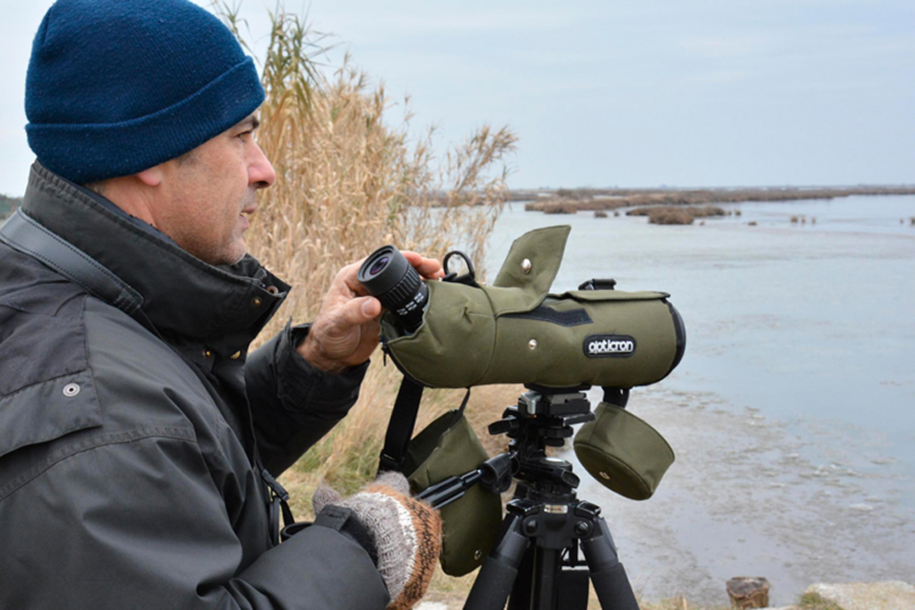 Foto von Mauro Bon. Er steht vor einem Fernglas auf einem Stativ an einem See.