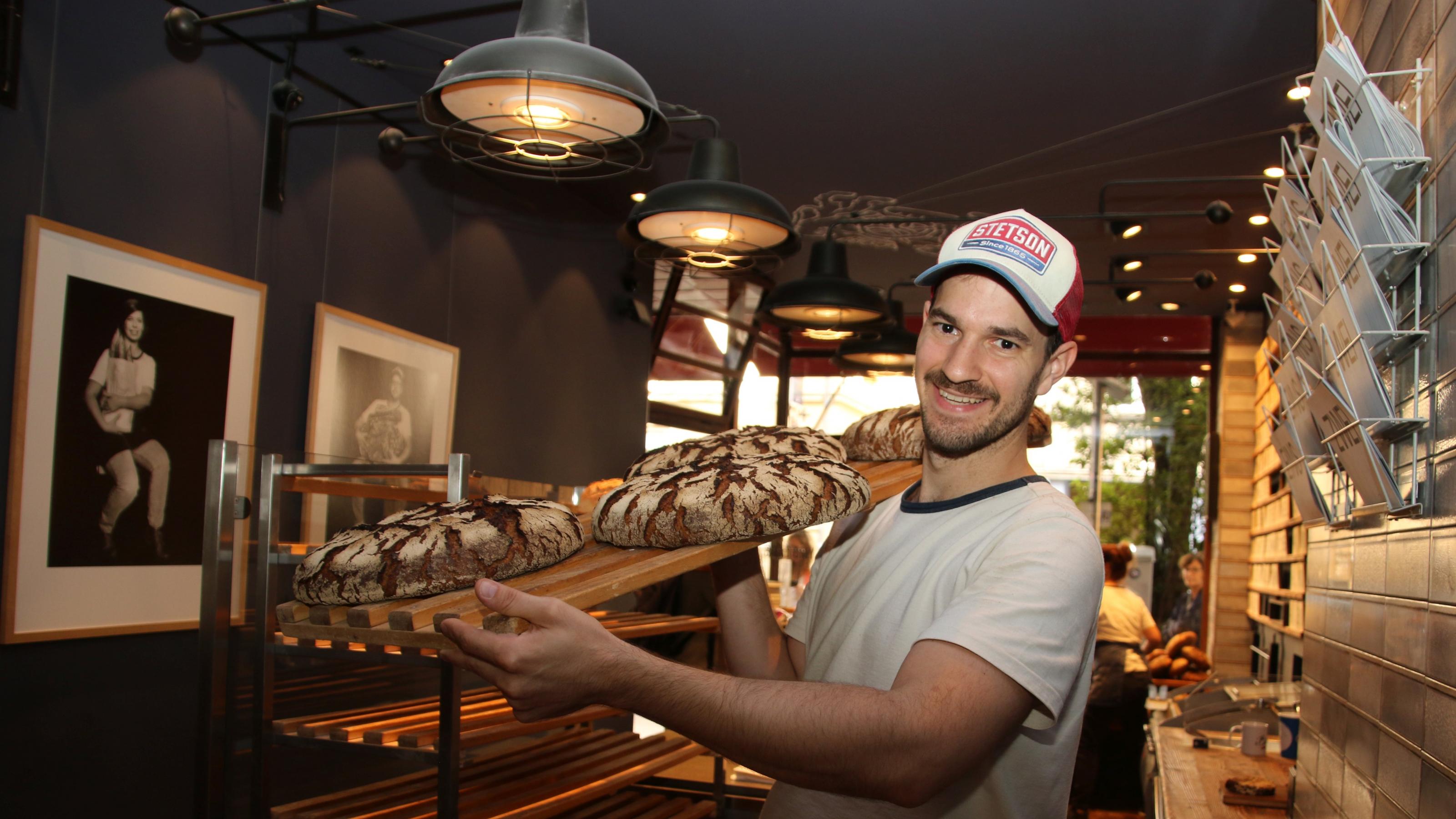 Der Bäcker Max Kugel trägt ein Holzbrett mit vier Broten auf der Schulter.