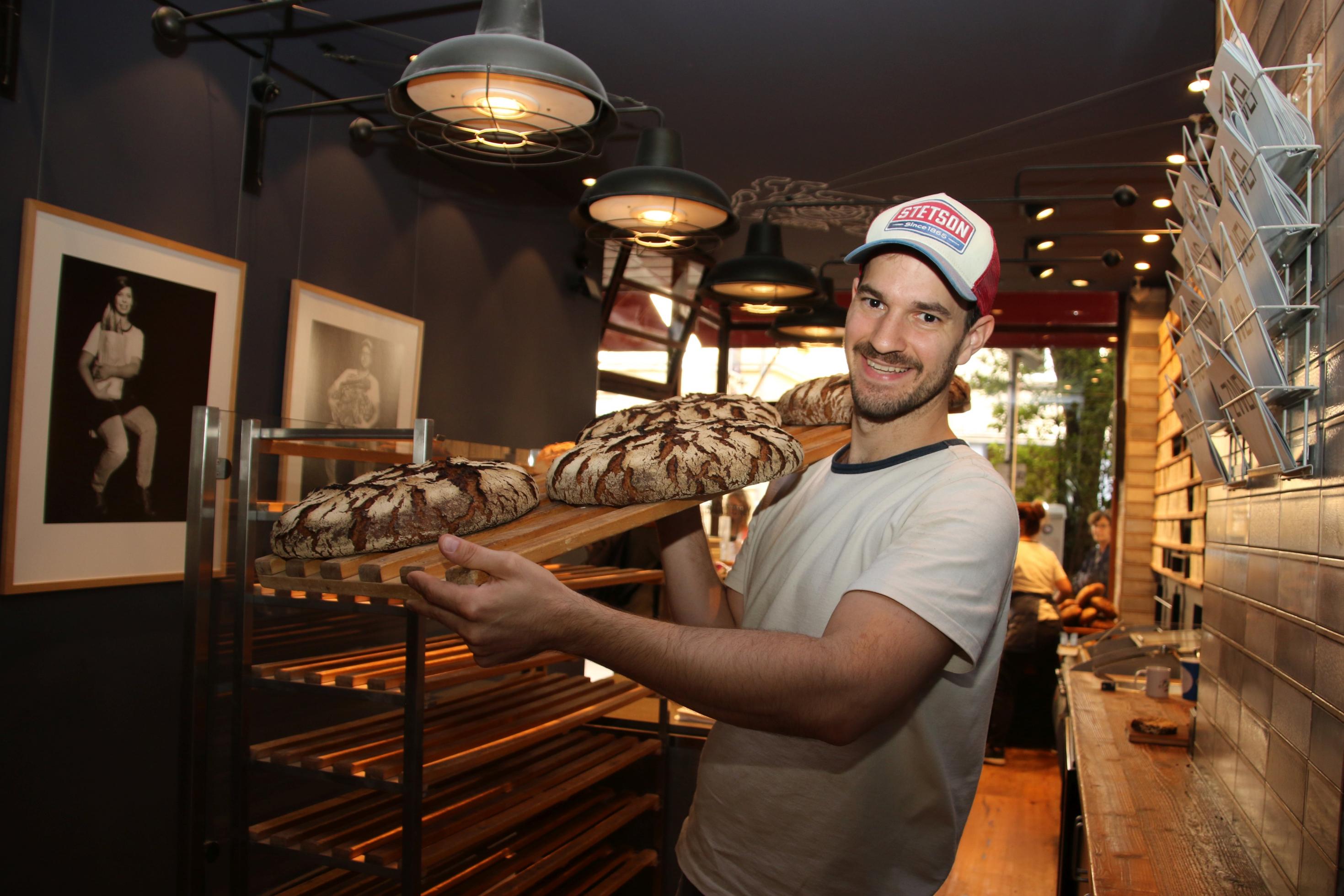 Der Bäcker Max Kugel trägt ein Holzbrett mit vier Broten auf der Schulter.