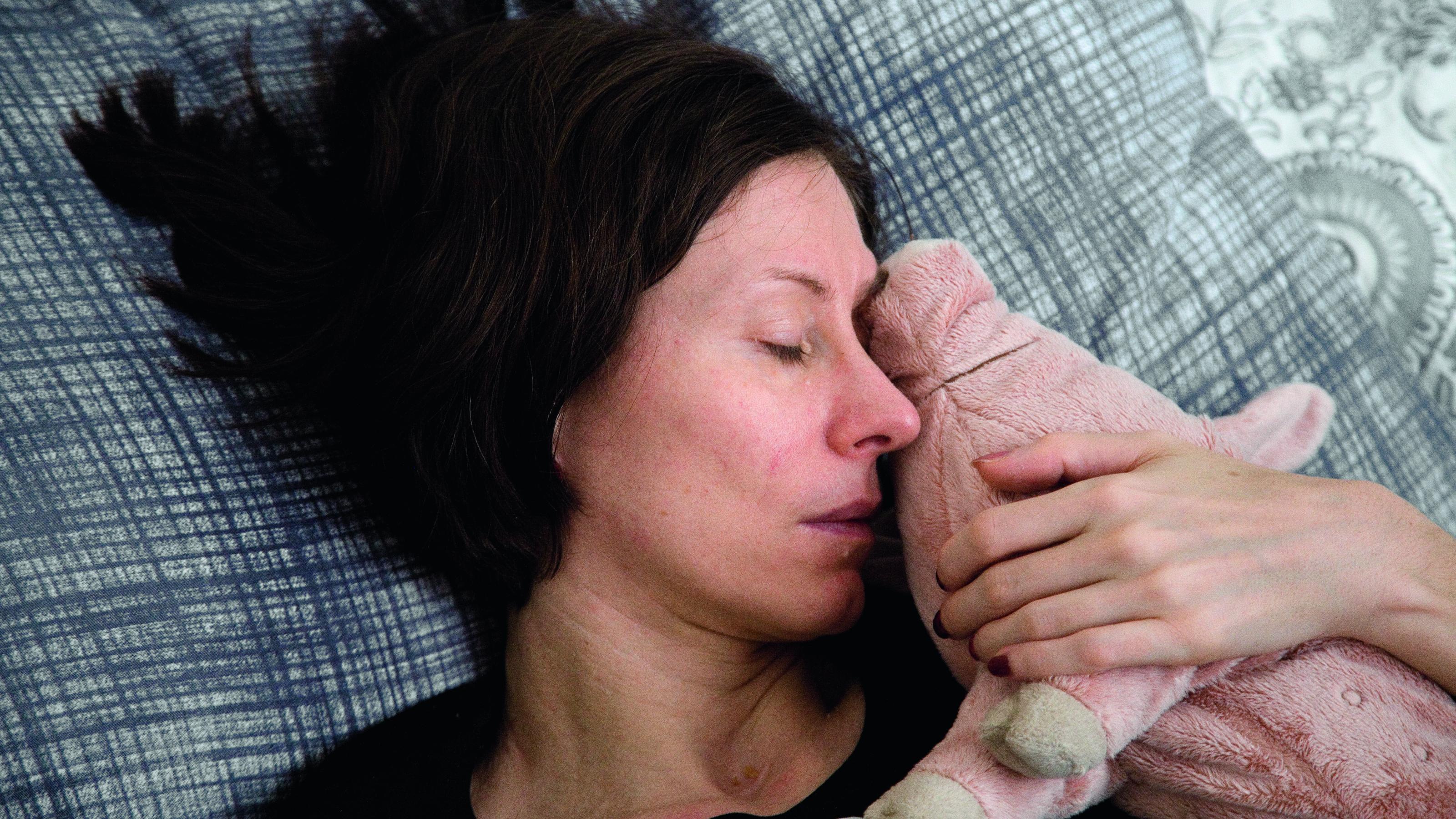 Eine erschöpft aussehende, an ME/CFS erkrankte Frau liegt in ihrem Bett, im Arm ein rosafarbenes Plüsch-Schwein.