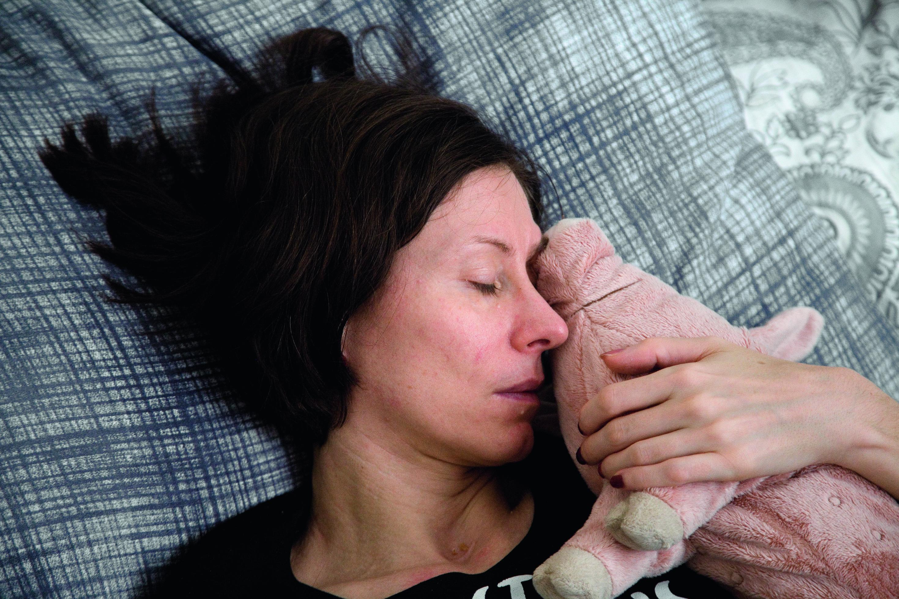 Eine erschöpft aussehende, an ME/CFS erkrankte Frau liegt in ihrem Bett, im Arm ein rosafarbenes Plüsch-Schwein.