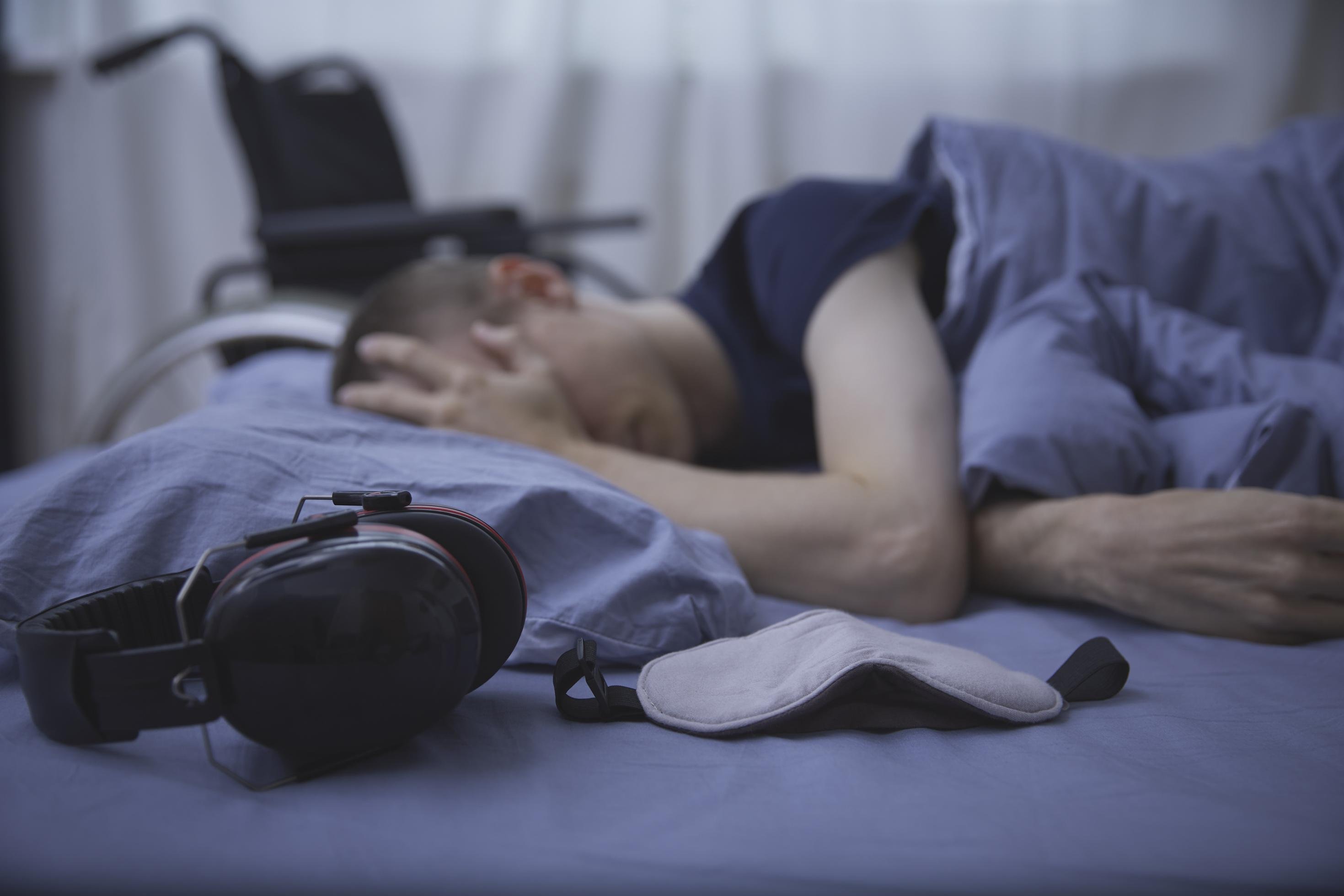 Schwer kranke Person liegt in einem Bett und hält eine Hand an die Stirn. Auf der Matratze vor ihr liegen Kopfhörer und eine Augenmaske, im Bildhintergrund ist ein Rollstuhl erkennbar.
