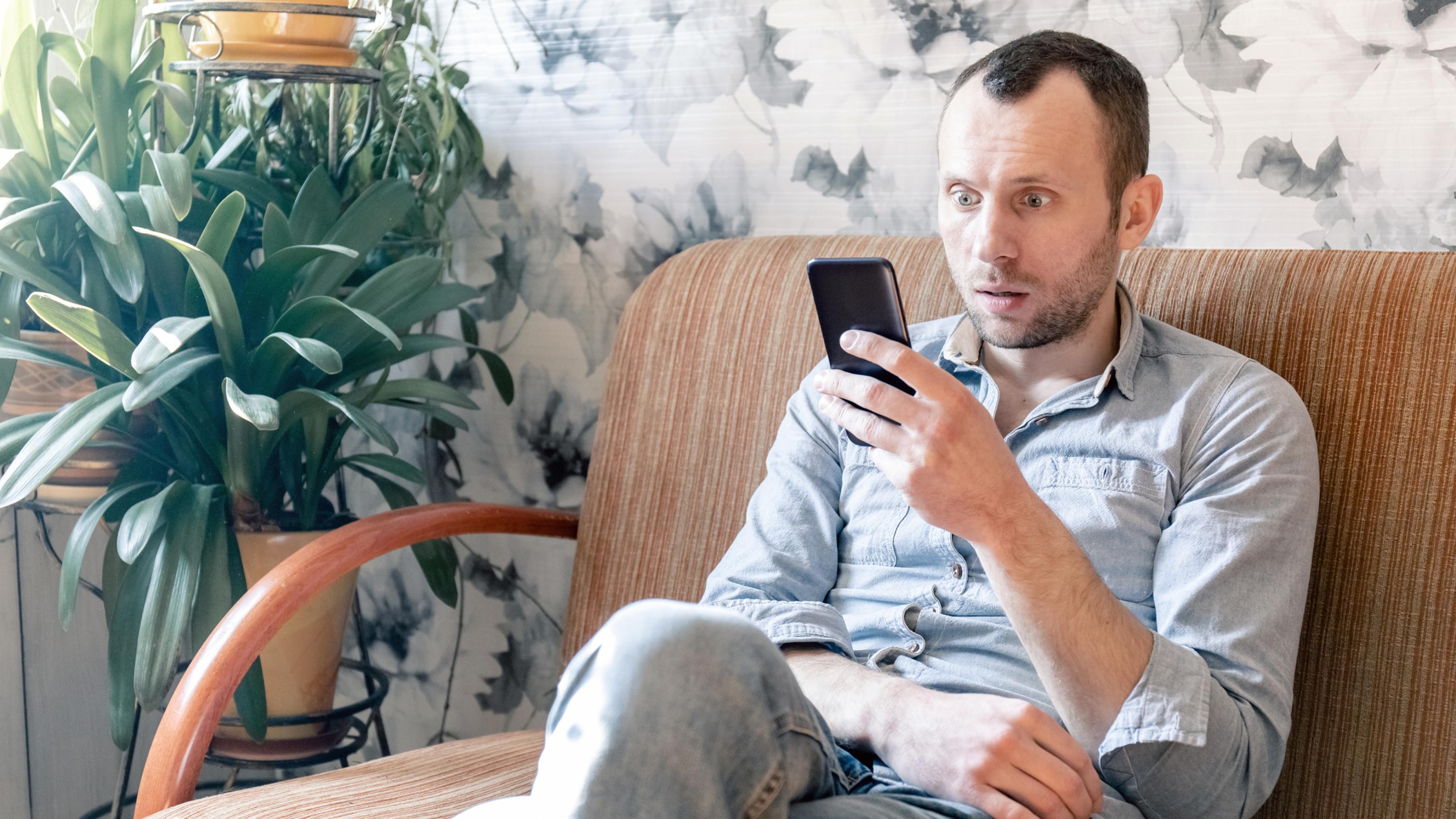 Ein junger Mann sitzt mit Angst im Gesicht während der Covid-19 Pandemie auf einem Sofa und liest Nachrichten auf seinem Smartphone.