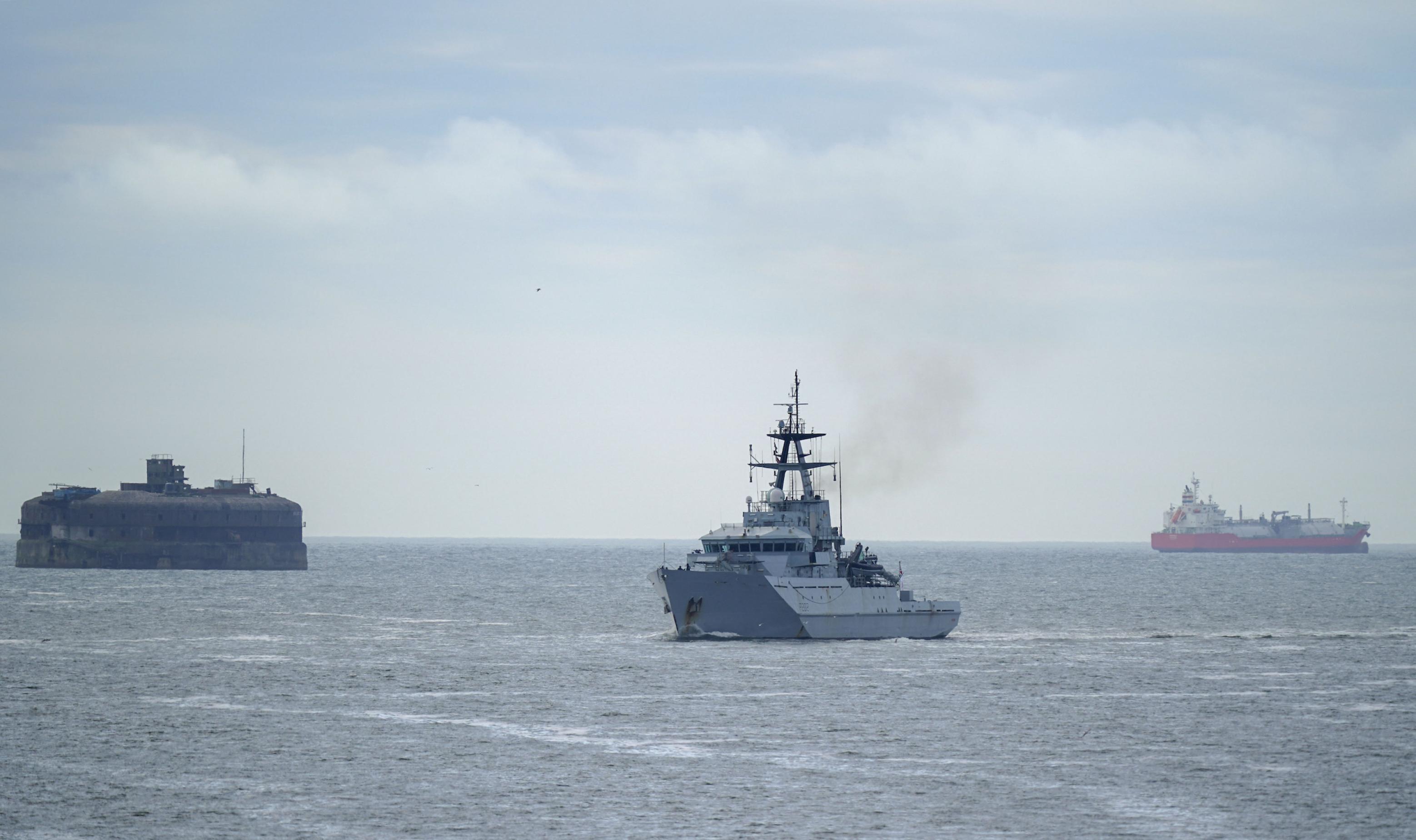 Mittelgroßes Kriegsschiff in grau auf grauer Meeresfläche, im Hintergrund größere zivile Schiffe.