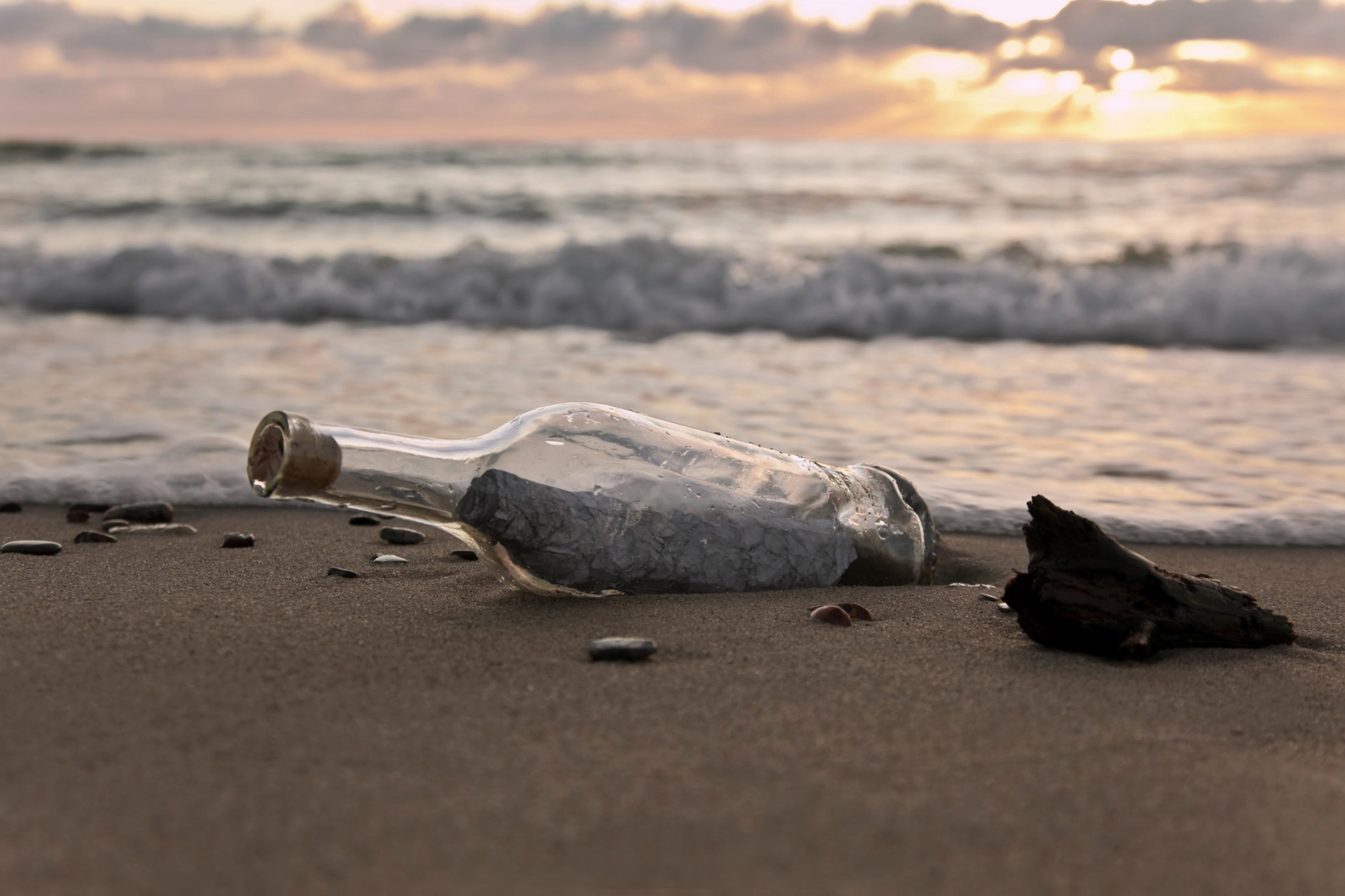 Eine durchsichtige Flasche mit einem Stück Papier drin liegt am Strand, dahinter Wellen und Dämmerungslicht