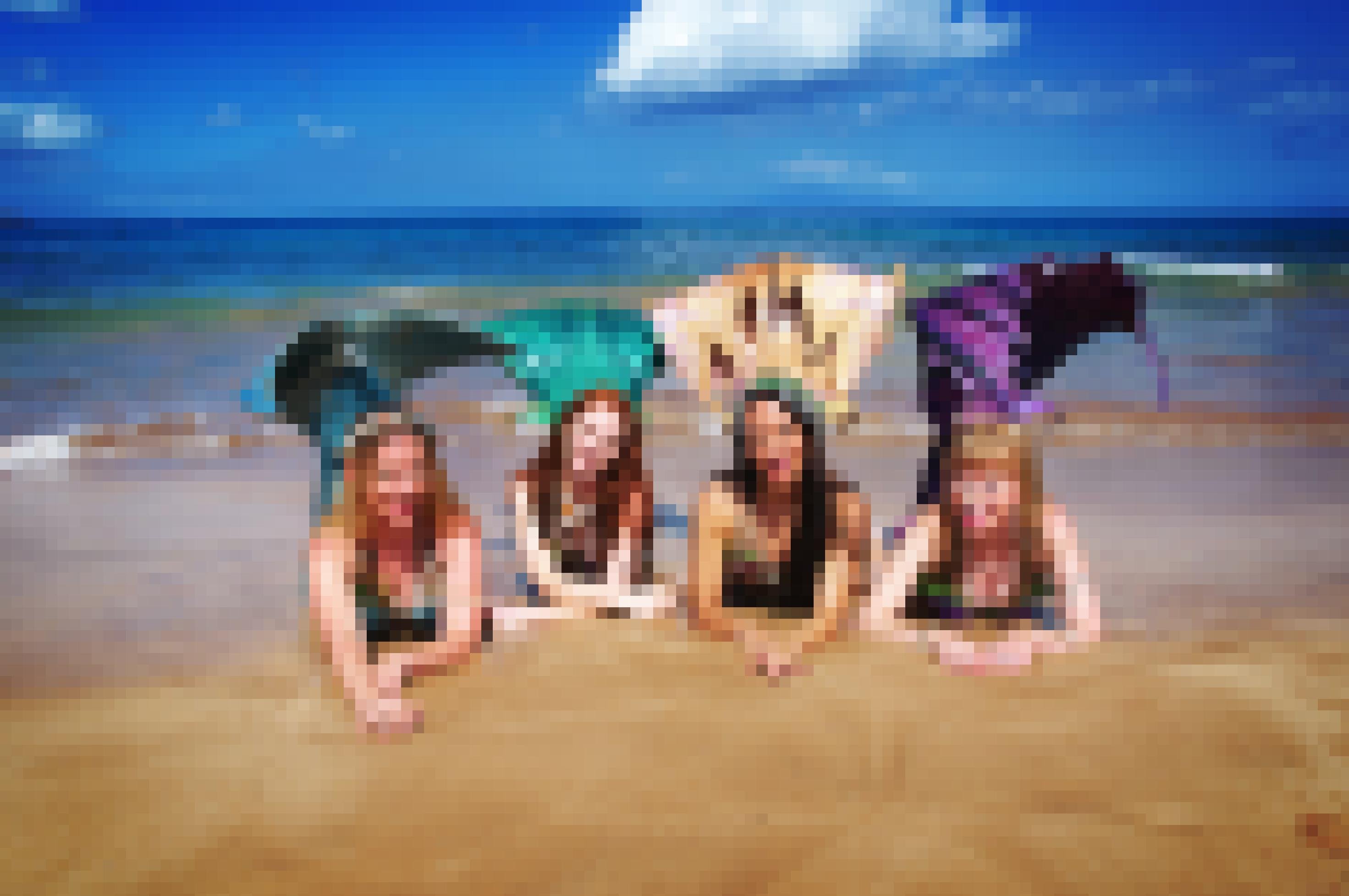 vier Frauen posieren in Meerjungfrauen-Kostümen am Meer, zwei blond, eine rothaarig, eine dunkle Haare. Sie liegen auf dem Bauch am Strand, die grünen, goldenen und lila Schwanzflossen angewinkelt und hochgestellt.