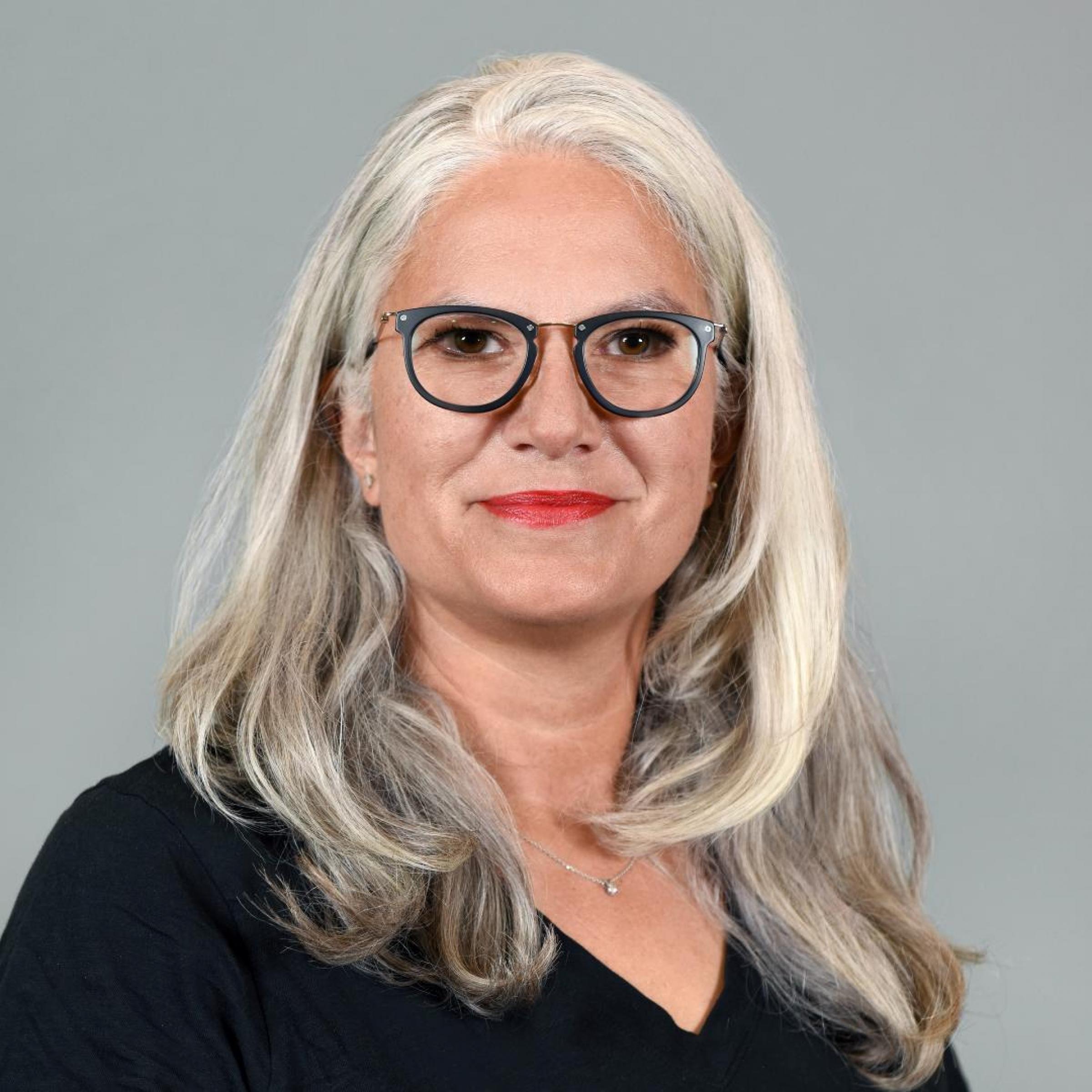 Porträtfoto von weißer Frau mit überschulterlangen grauen Haaren. Sie trägt eine Brille mit schwarzem Gestell und lächelt.