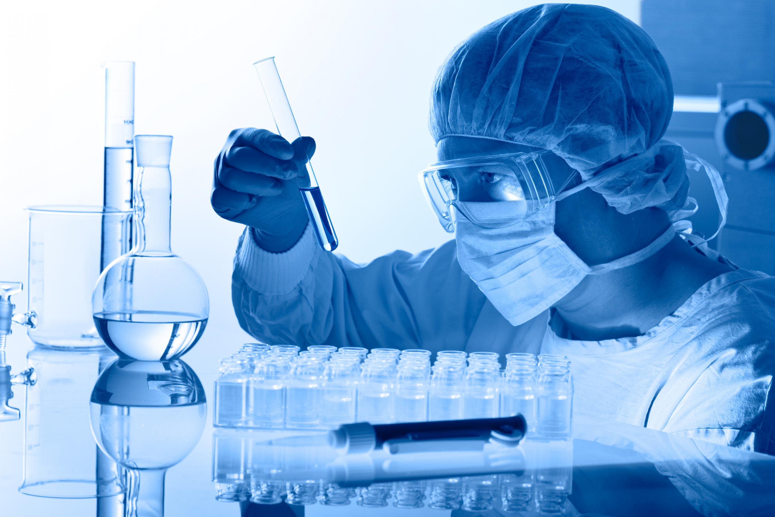 Eine Person mit Mundschutz und Schutzbrille schaut in einem Labor in ein Reagenzglas.