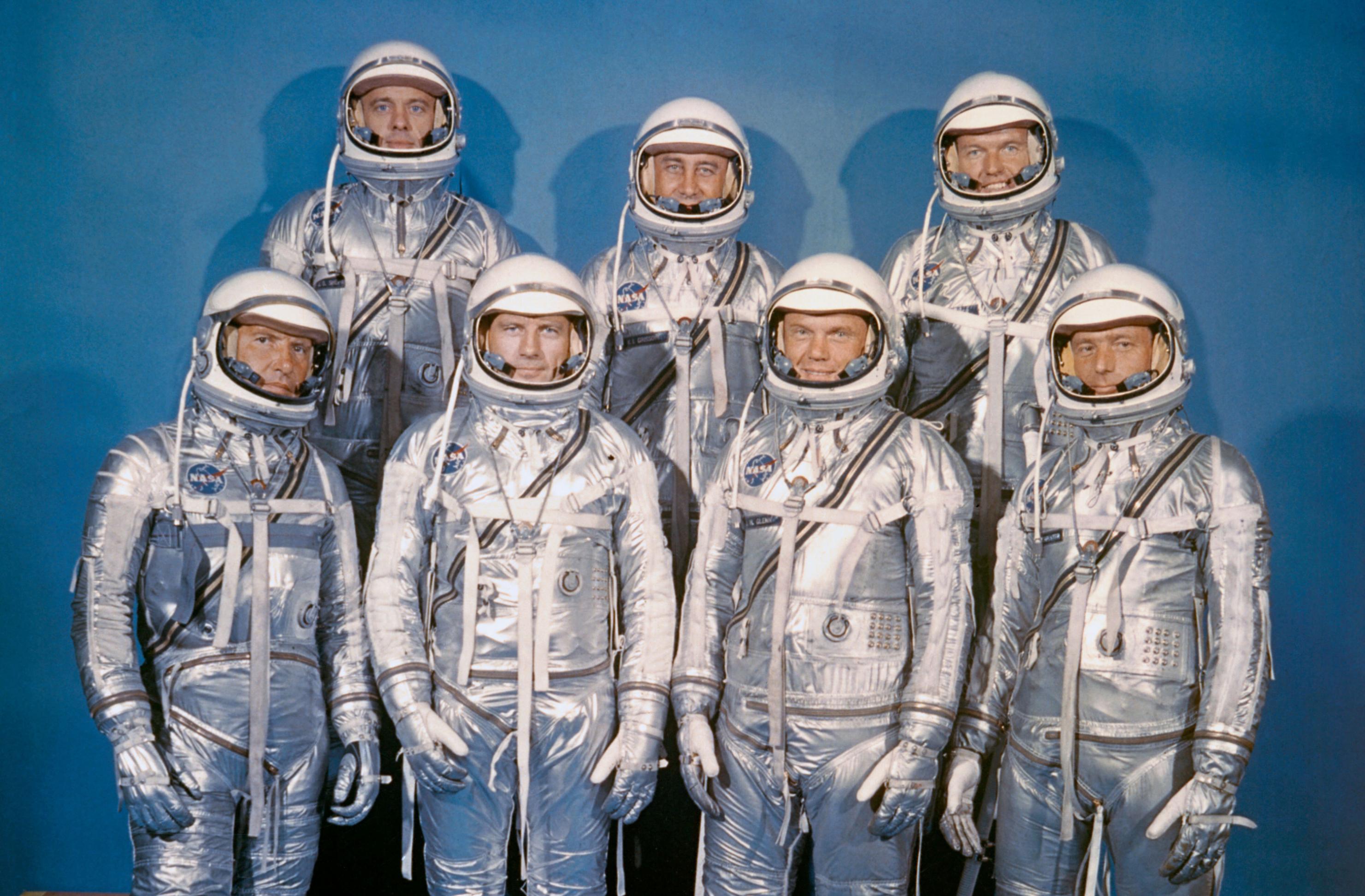Die Mercury 7, sieben Astronauten in silbrigen Anzügen posieren vor blauer Leinwand: Walter Schirra, Donald „Deke“ Slayton, John H. Glenn, Scott Carpenter; hintere Reihe: Alan B. Shepard, Virgil I. „Gus“ Grissom und Gordon Cooper