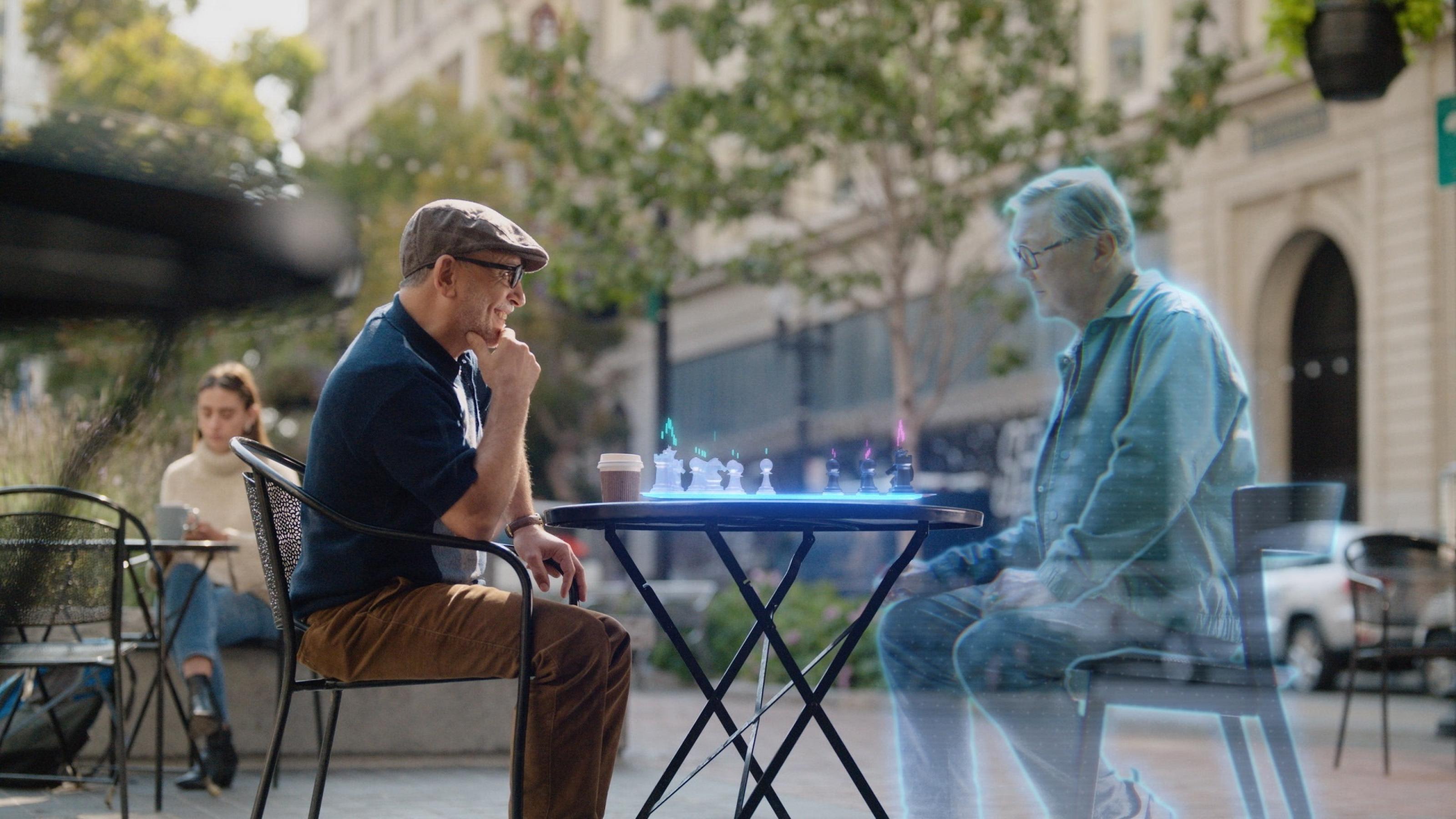 Ein älterer Mann sitzt auf einem realen Platz an einem Tisch mit einem digitalen Schachbrett, ein digitaler Schachpartner wird im Metaverse eingeblendet.