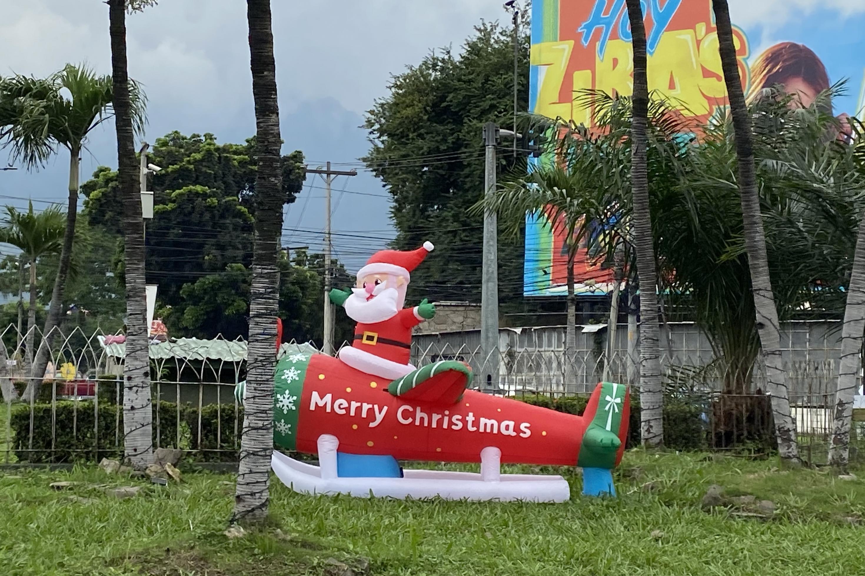 In einem Garten oder Park mit Palmen steht auf dem Rasen eine aufblasbarer Rakete mit Kufen. Auf dem Ding reitet mit ausgebreiteten Armen ein Gummiweihnachtsmann. Über sein Gefährt steht quer geschrieben: Merry Christmas.