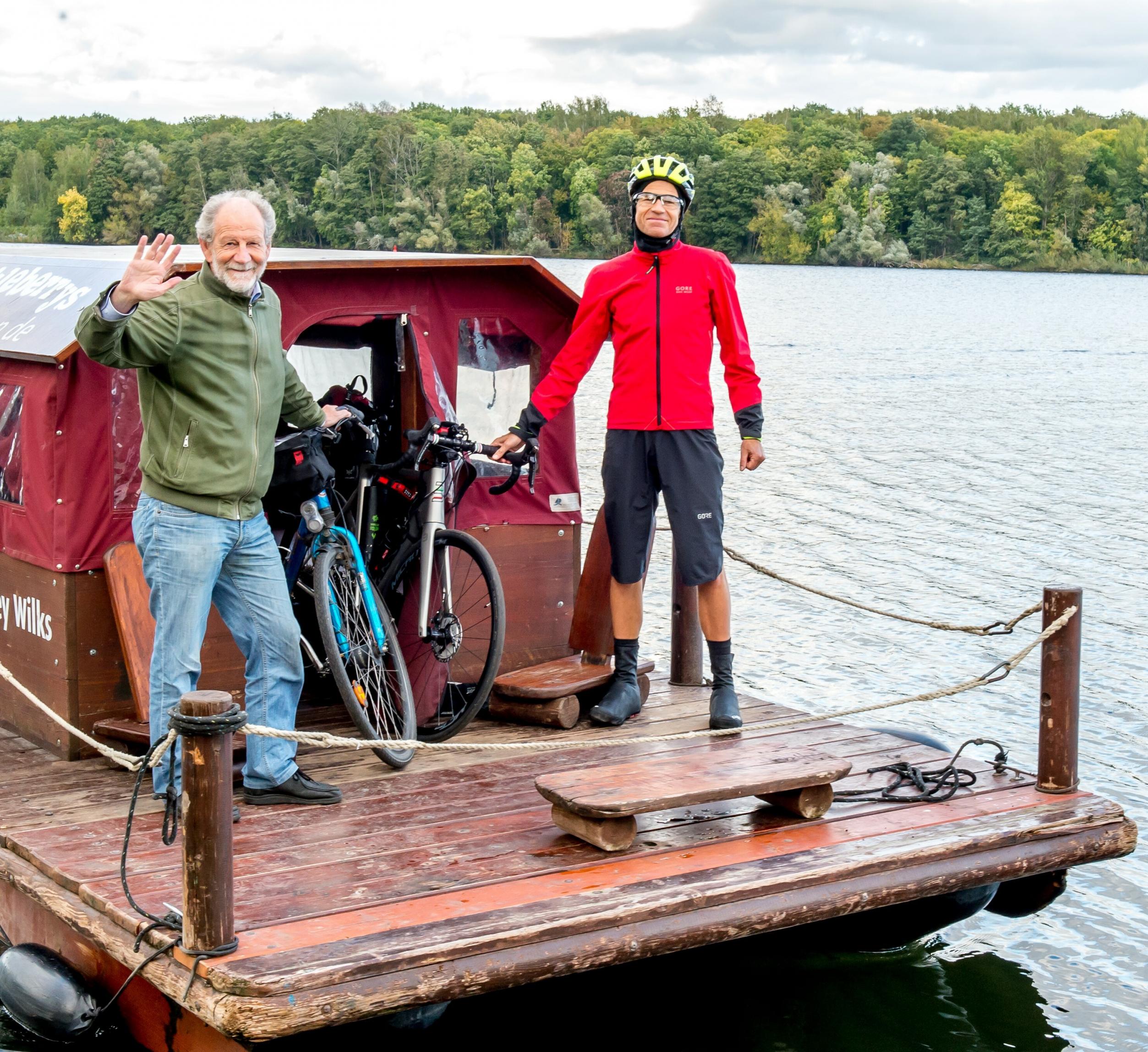 Die beiden Männer halten mit jeweils einer Hand ihr Fahrrad fest und stehen auf dunklen Holzplanken eines Floßes. Im Hintergrund ist der Jungfernsee und der Herbstwald des Sacrower Nordufers zu sehen.