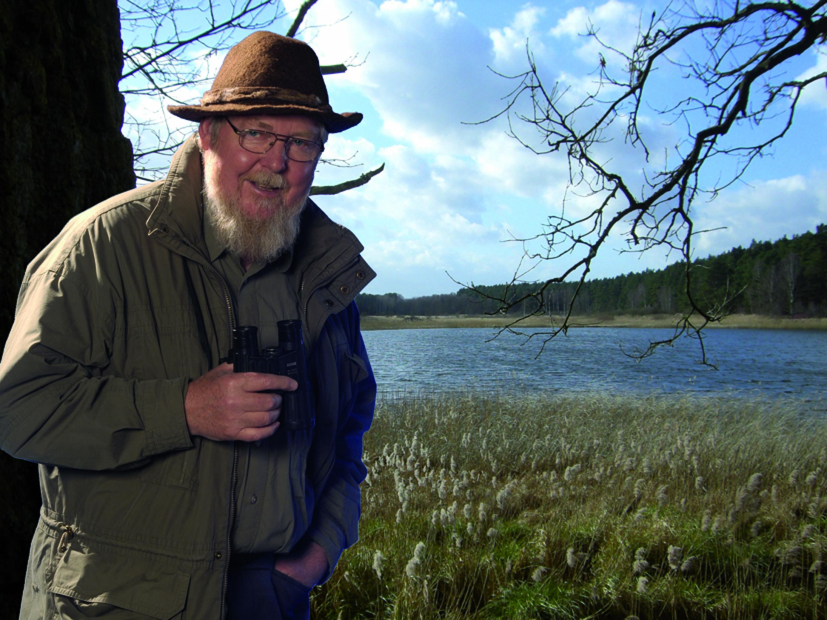 Mann mit Vollbart, Hut und Fernglas in Landschaft mit Wasser