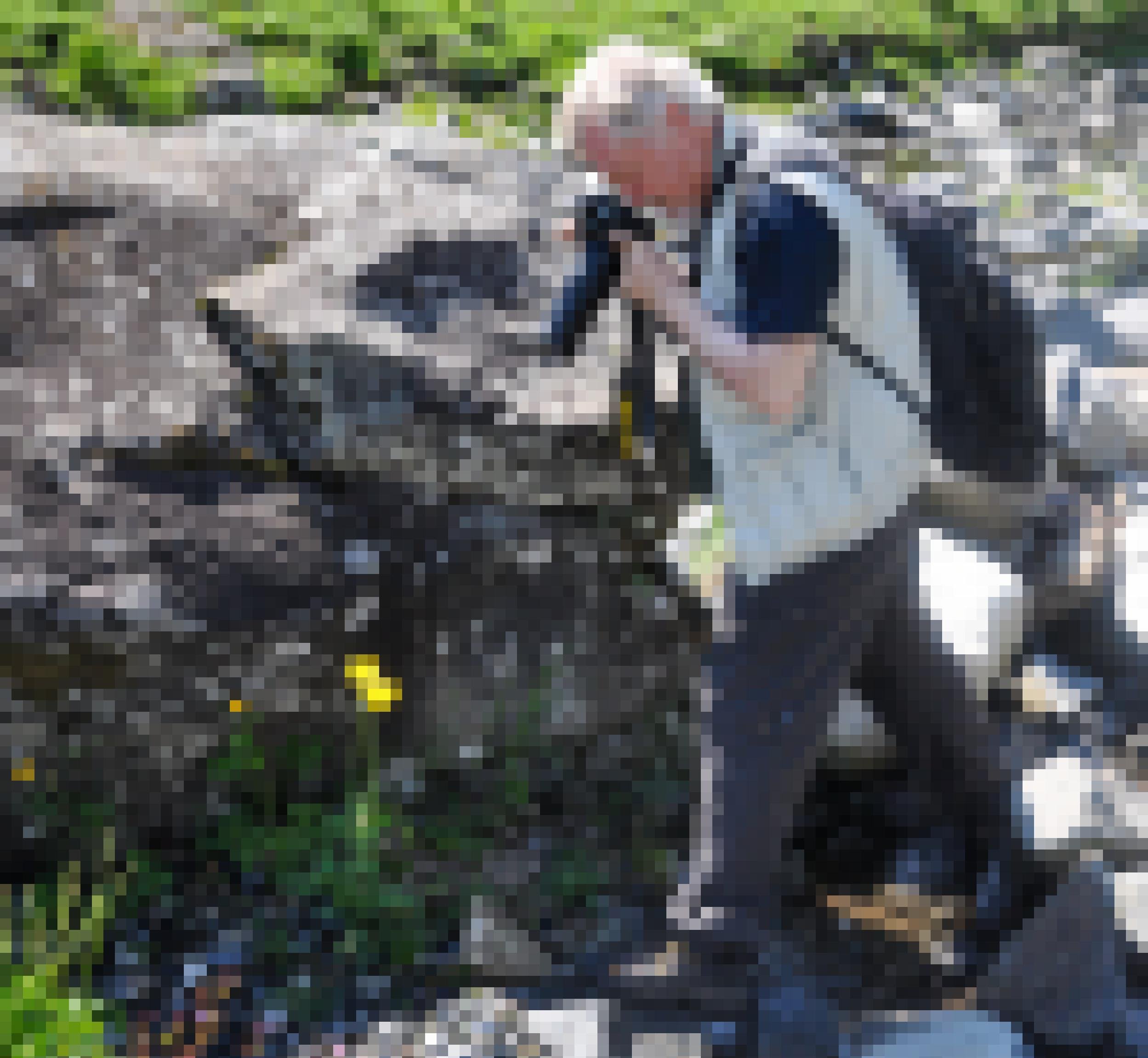 Ein Mann mit weißen Haaren und Bart fotografiert mit einem langen Objektiv eine gelbe Blüte in einer Felslandschaft.