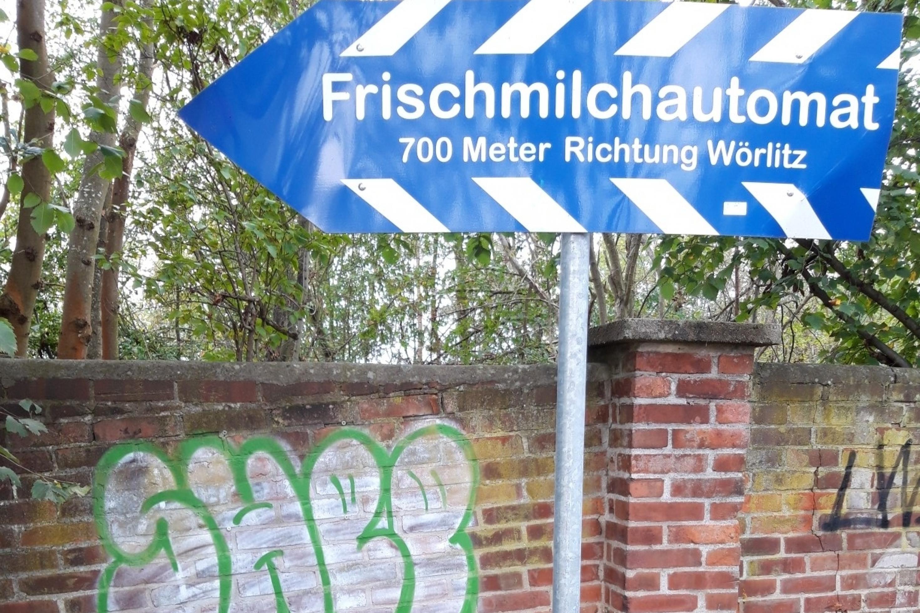 Das Rennrad des RadelndenReporters lehnt am Pfahl eines Hinweisschilds, auf dem zu lesen ist „Frischmilchautomat – 700 Meter Richtung Wörlitz“.