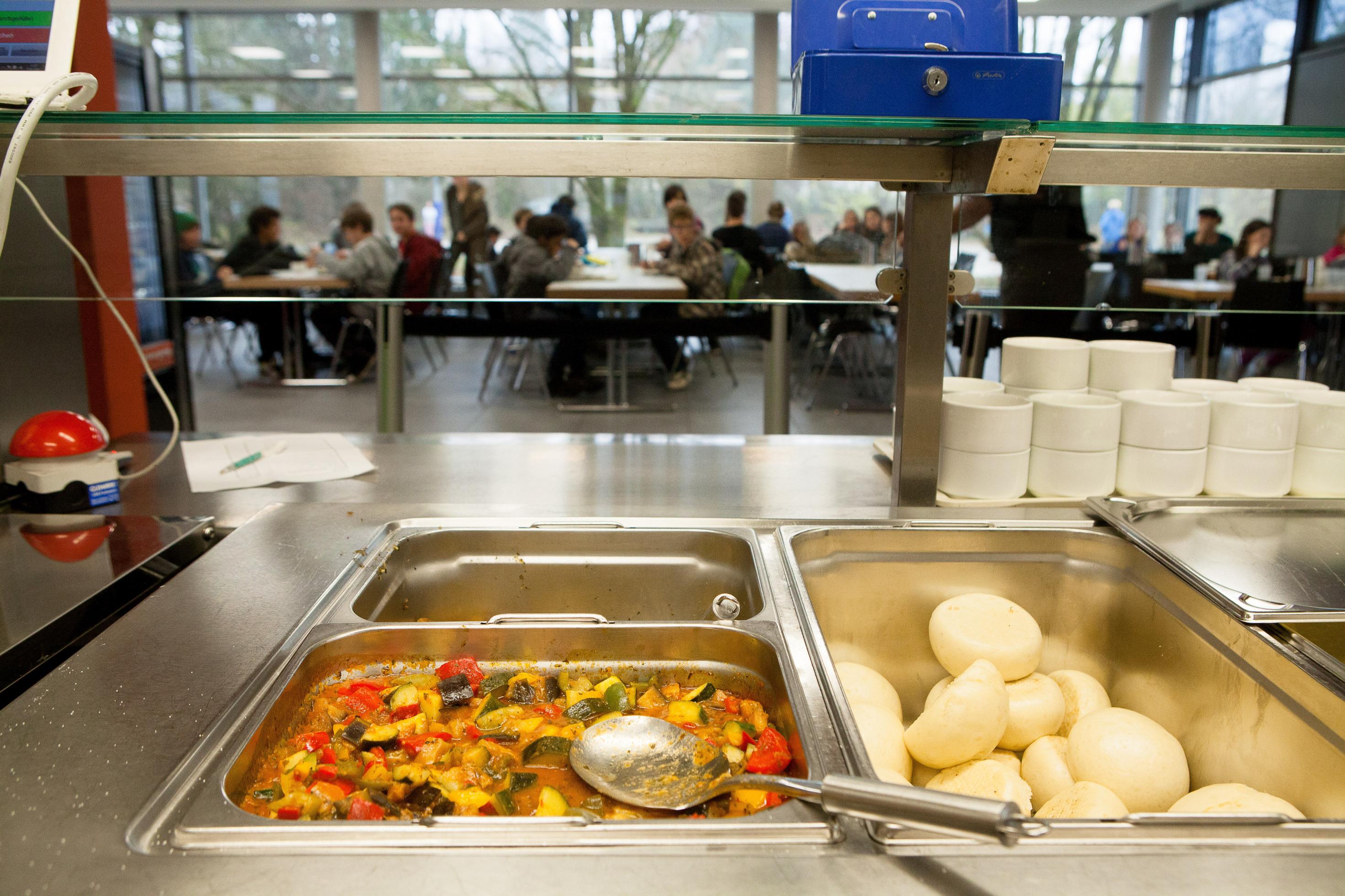 Die Essenausgabe einer Schulmensa. Im Vordergrund sieht man Ratatouille und Knödel in Edelstahlbehältern, im Hintergrund Kinder beim Mittagessen an langen Tischen.
