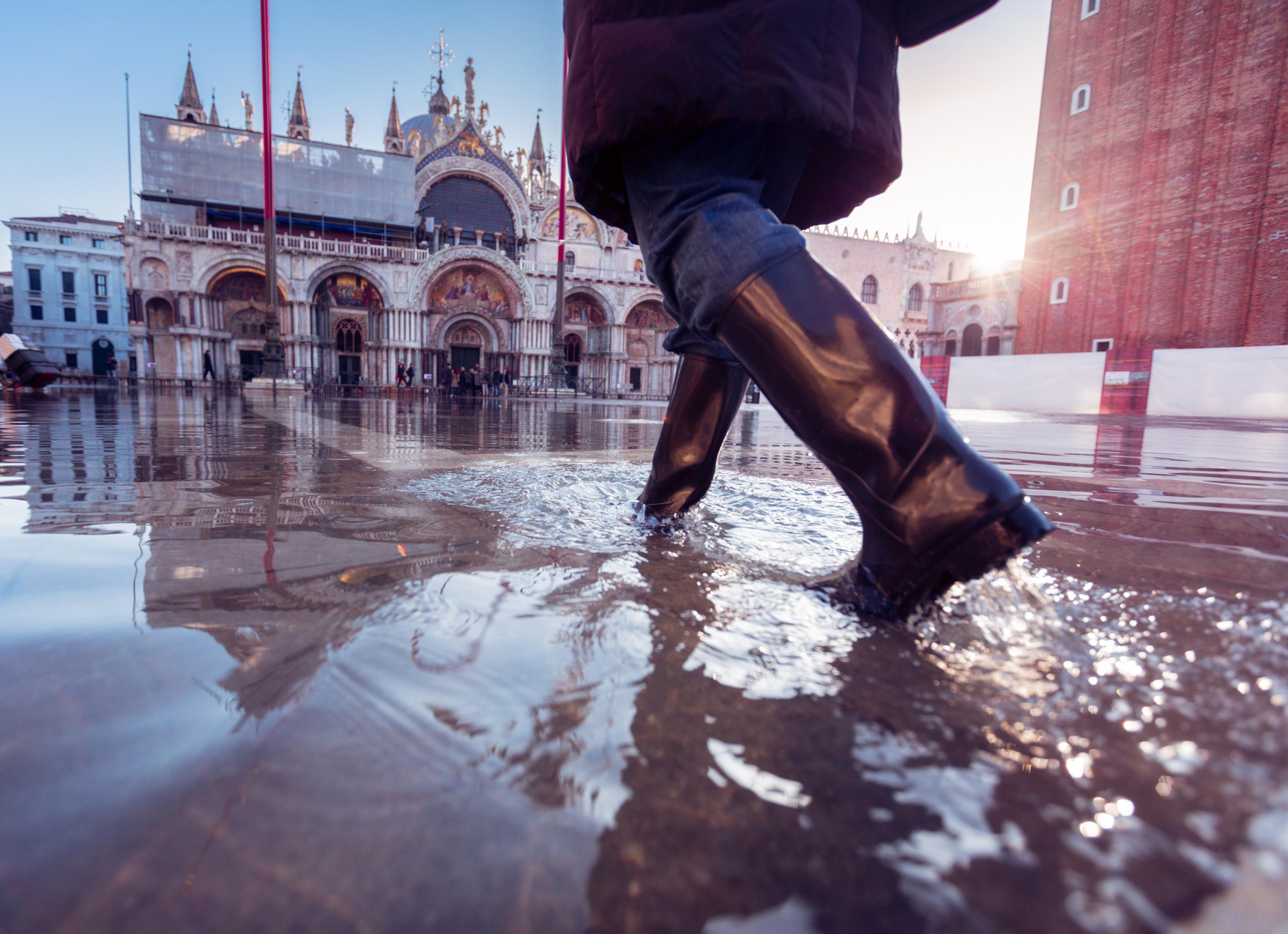 Frau in Gummistiefeln watet am Markusplatz durch Wasser.