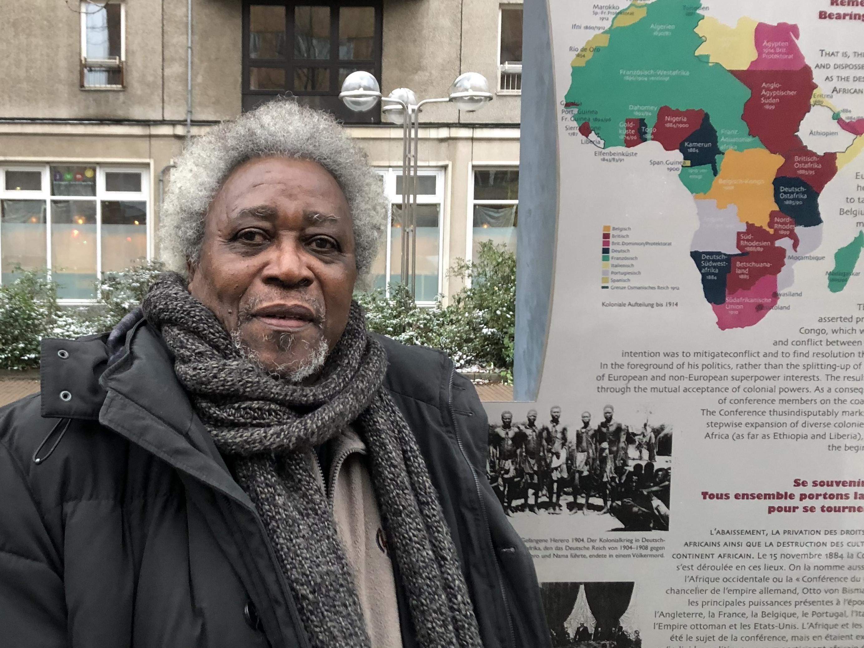 Ein älterer Mann mit grauem Haar vor einer Infotafel mit Informationen zum Kolonialismus.