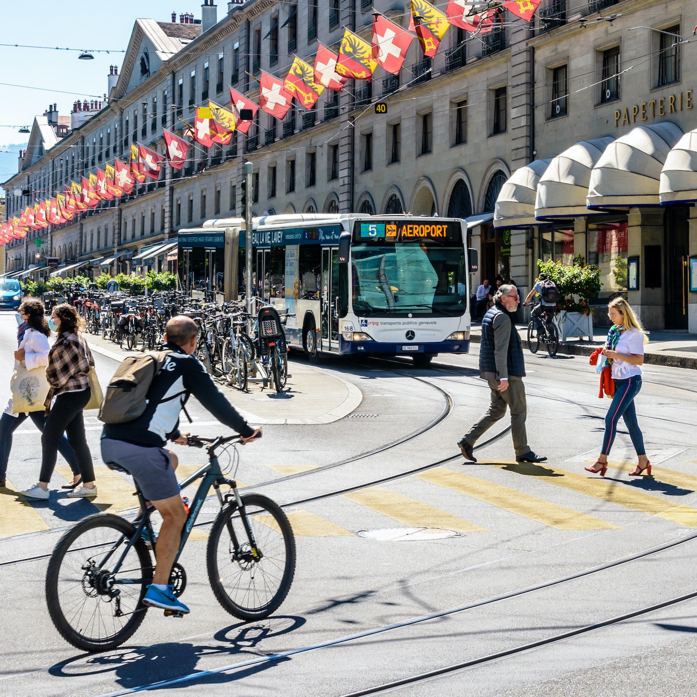 Genf, Schweiz: Fußgänger und Radfahrer überqueren die Straße, ein Bus wartet an einer roten Ampel.