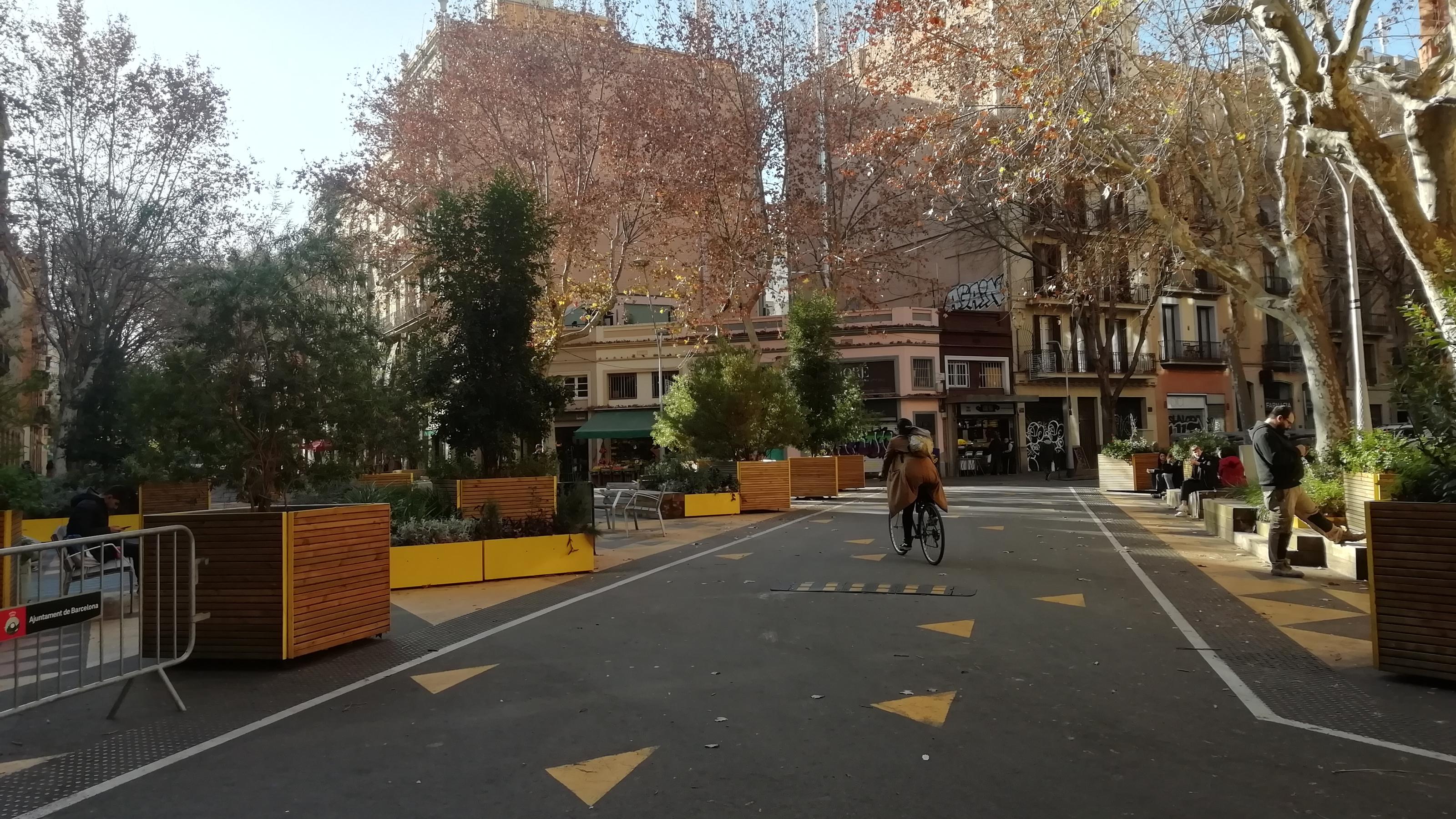 Blick auf die verkehrsberuhigte Superilla in Sant Antoni in Barcelona. Die Mitte der Straßenkreuzung ist mit Pflanzenkübeln für den Verkehr blockiert, drumherum stehen Holzbänke. Auf dem Boden sind gelbe Markierungen. Ein Radfahrer fährt über die Straße.