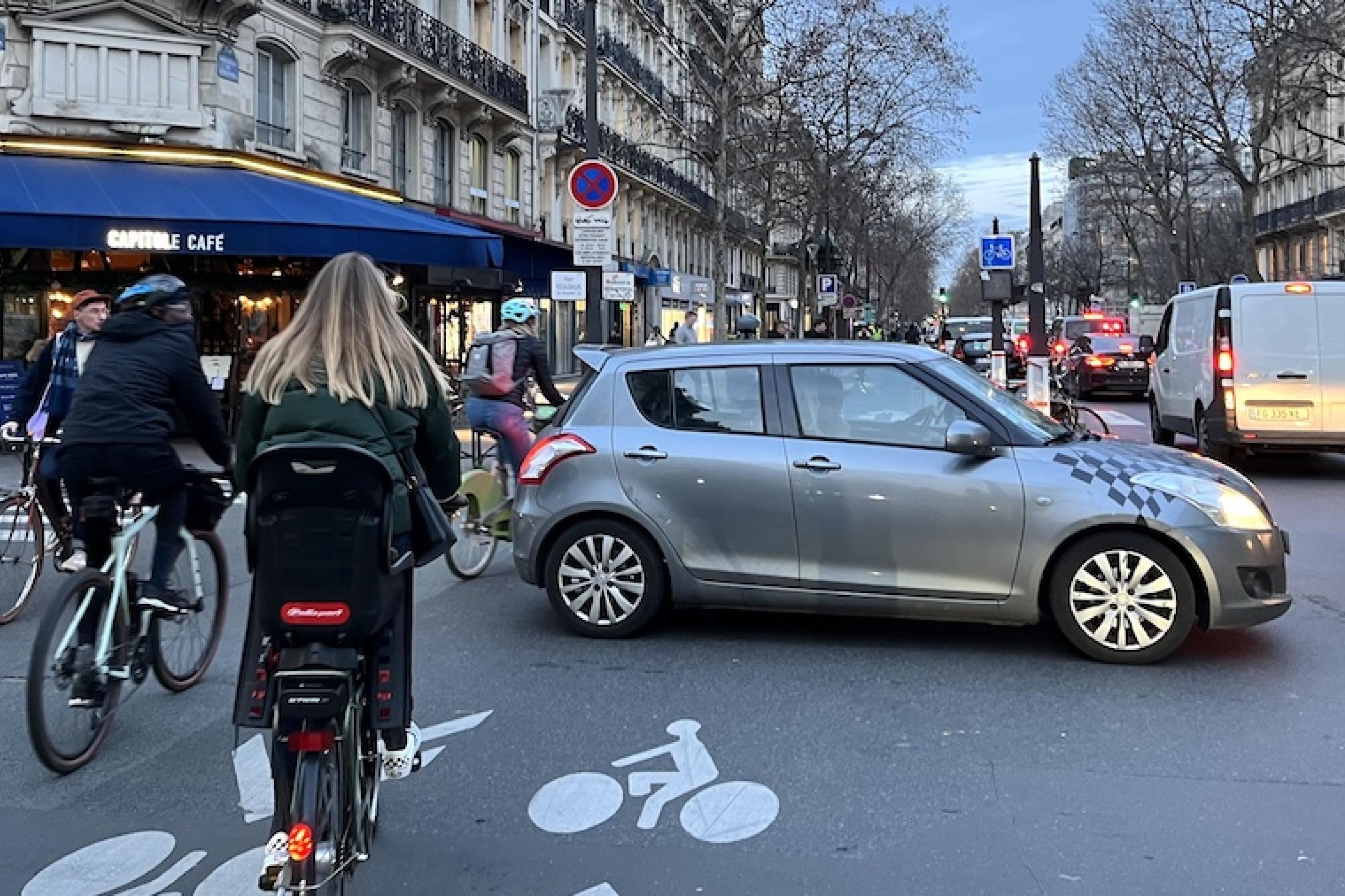 Eine Straßenszene in Paris. Auf der rechten Bildhälfte stauen sich PKWs, auf der der linken Radfahrer. Der Radweg wird von einem abbiegenden Auto blockiert.