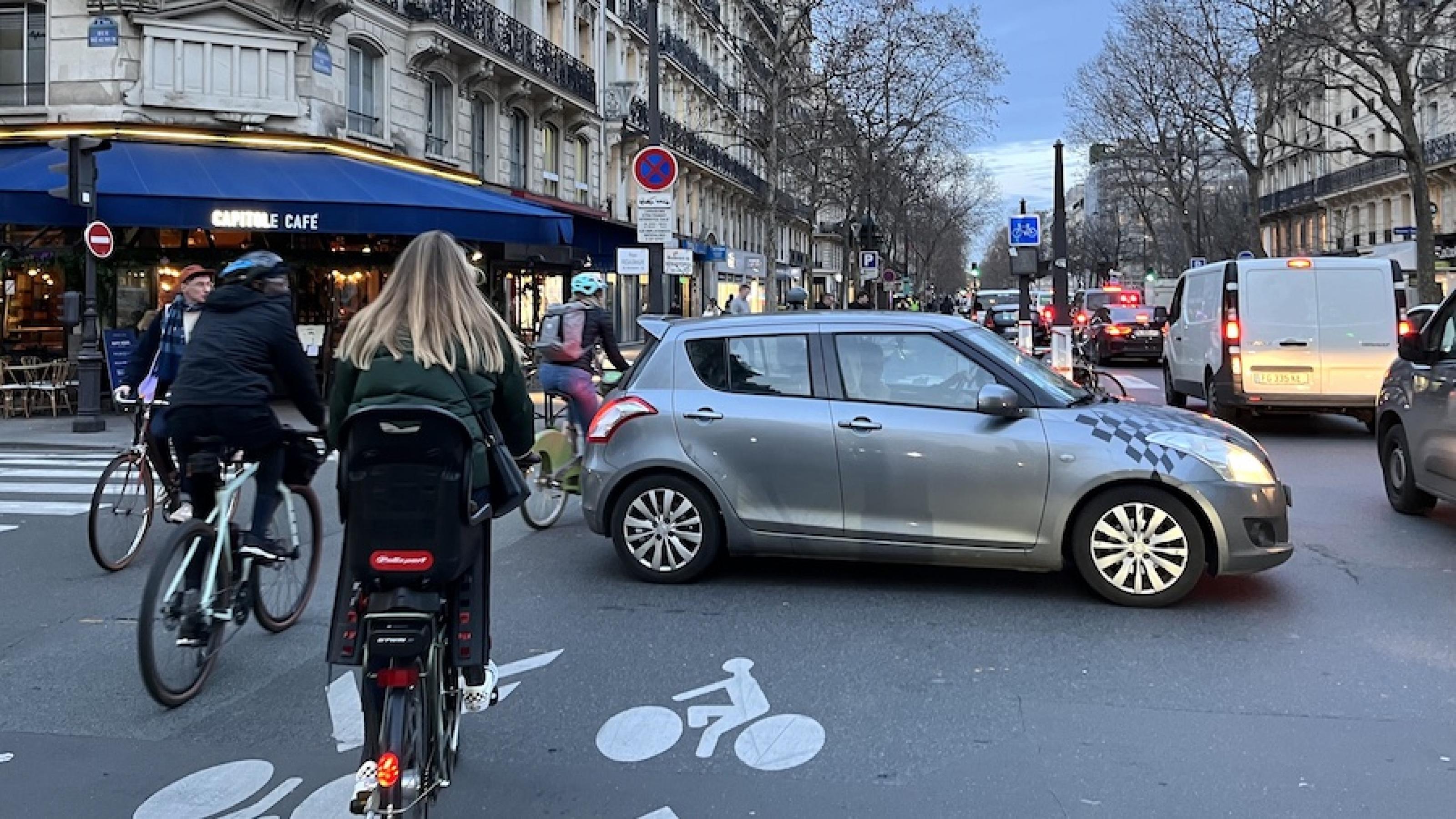 Eine Straßenszene in Paris. Auf der rechten Bildhälfte stauen sich PKWs, auf der der linken Radfahrer. Der Radweg wird von einem abbiegenden Auto blockiert.