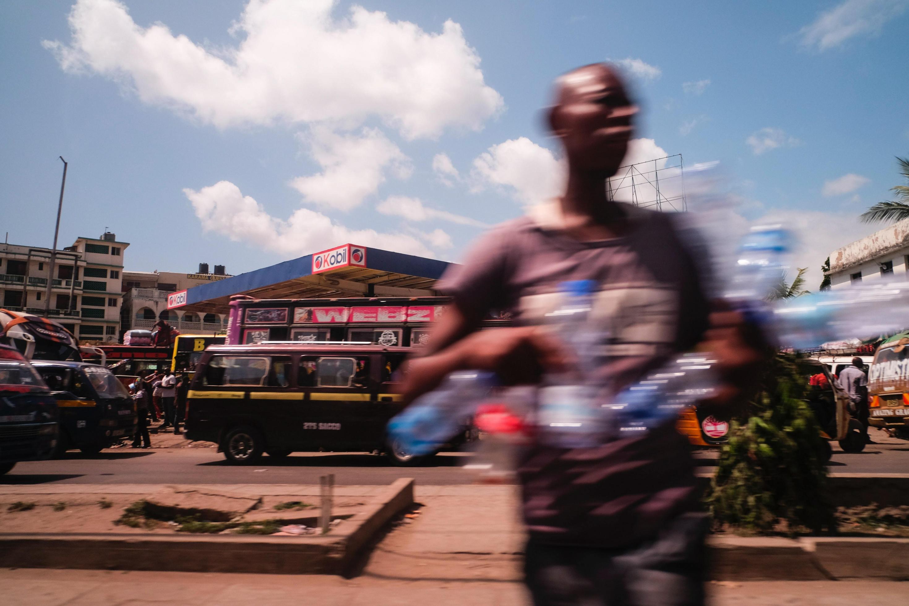Im Vordergrund steht durch die Bewegung verschwommen ein Mann, im Arm trägt er unterschiedliche Waren, die er auf der Straße zum Verkauf anbietet.