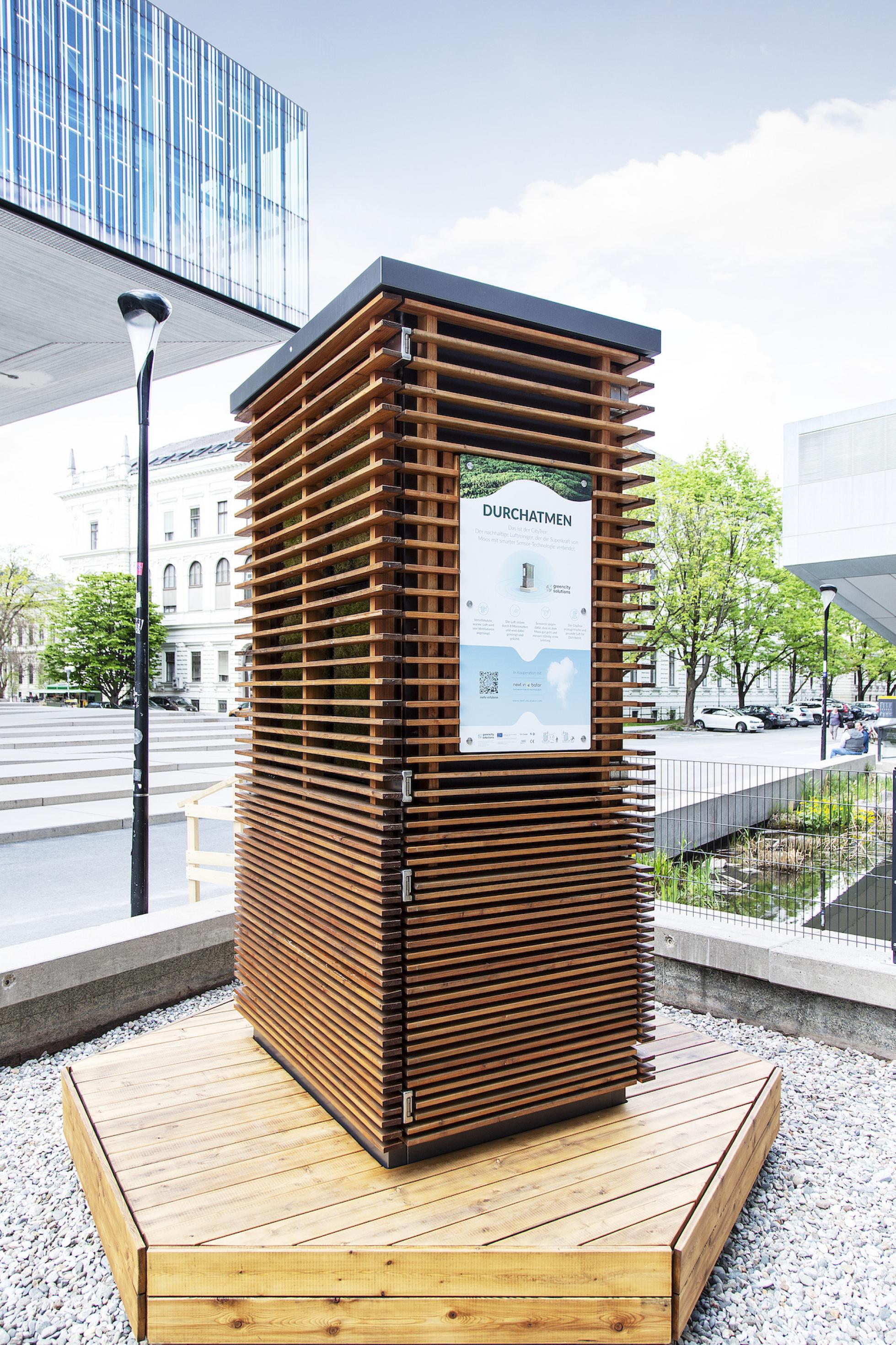 Viereckige, rund zwei Meter hohe Holzkonstruktion an einer Straße in Berlin. Auf der Vorderseite steht „Durchatmen“, am Fuß ist eine umlaufende Sitzbank angebracht.
