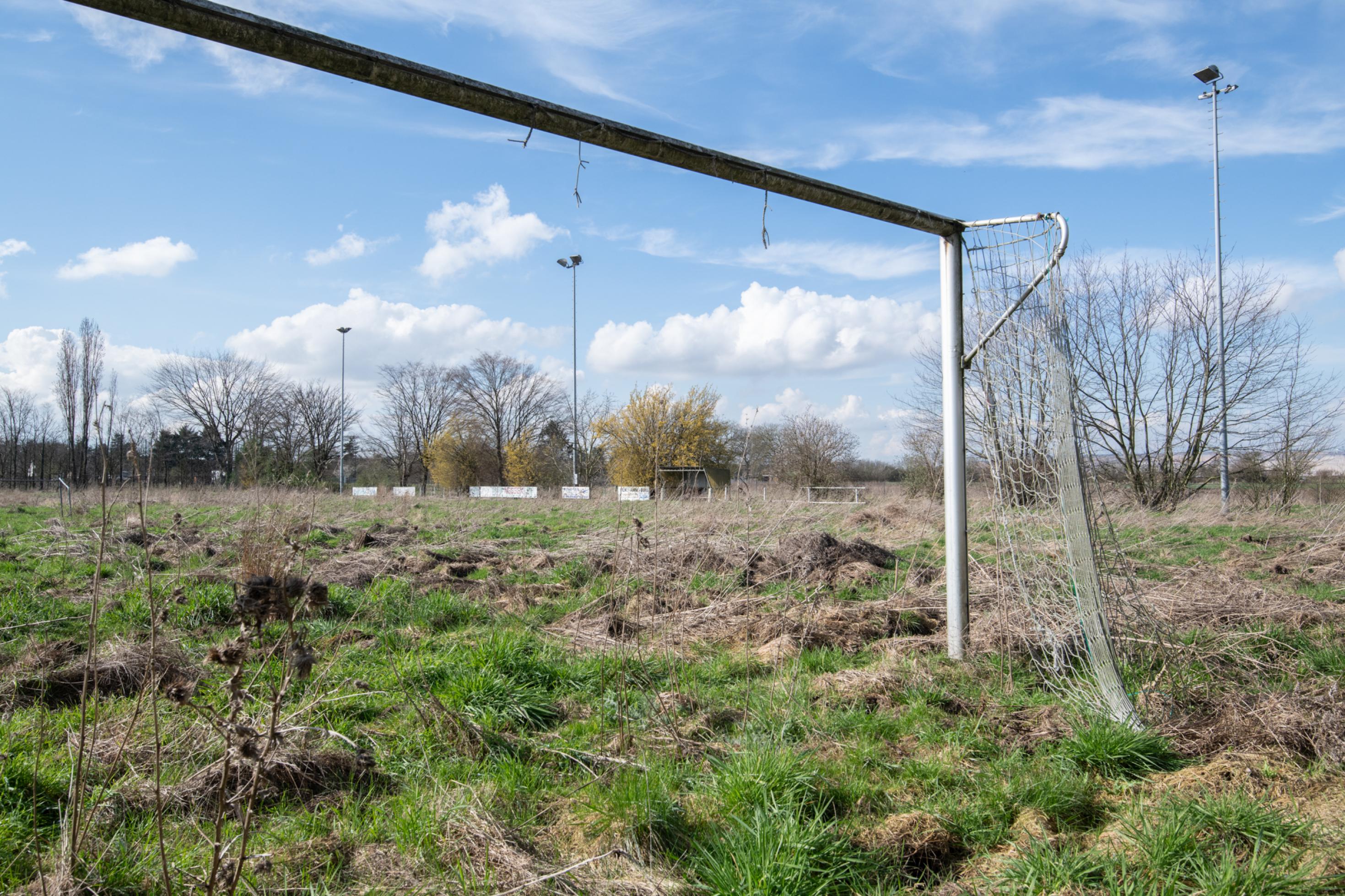 Blick durch ein kaputtes Fußbalaltor auf einen hoch mit Pflanzen bewachsenen einstigen Fußballplatz.