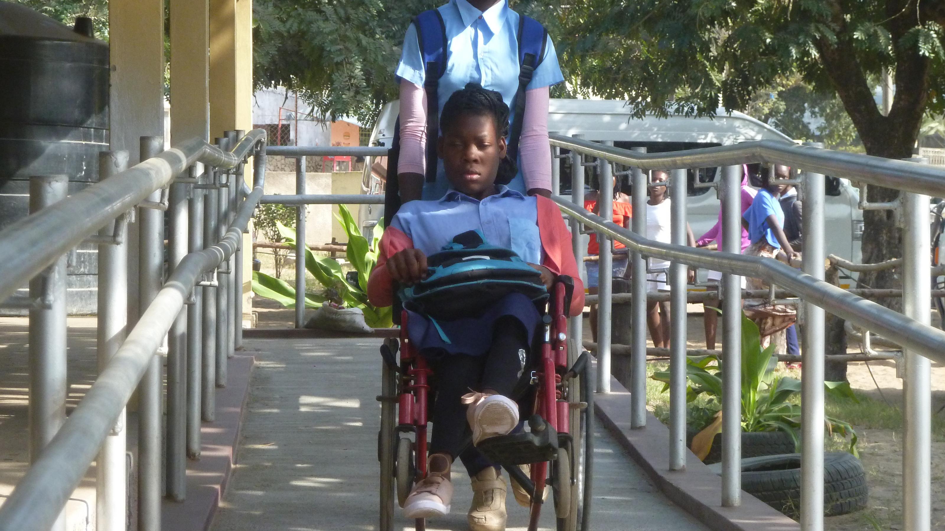 Chelsia sitzt im Rollstuhl, ihre Freundin schiebt sie die neue Rampe in der Schule hinunter, rechts und links ein Metallgeländer