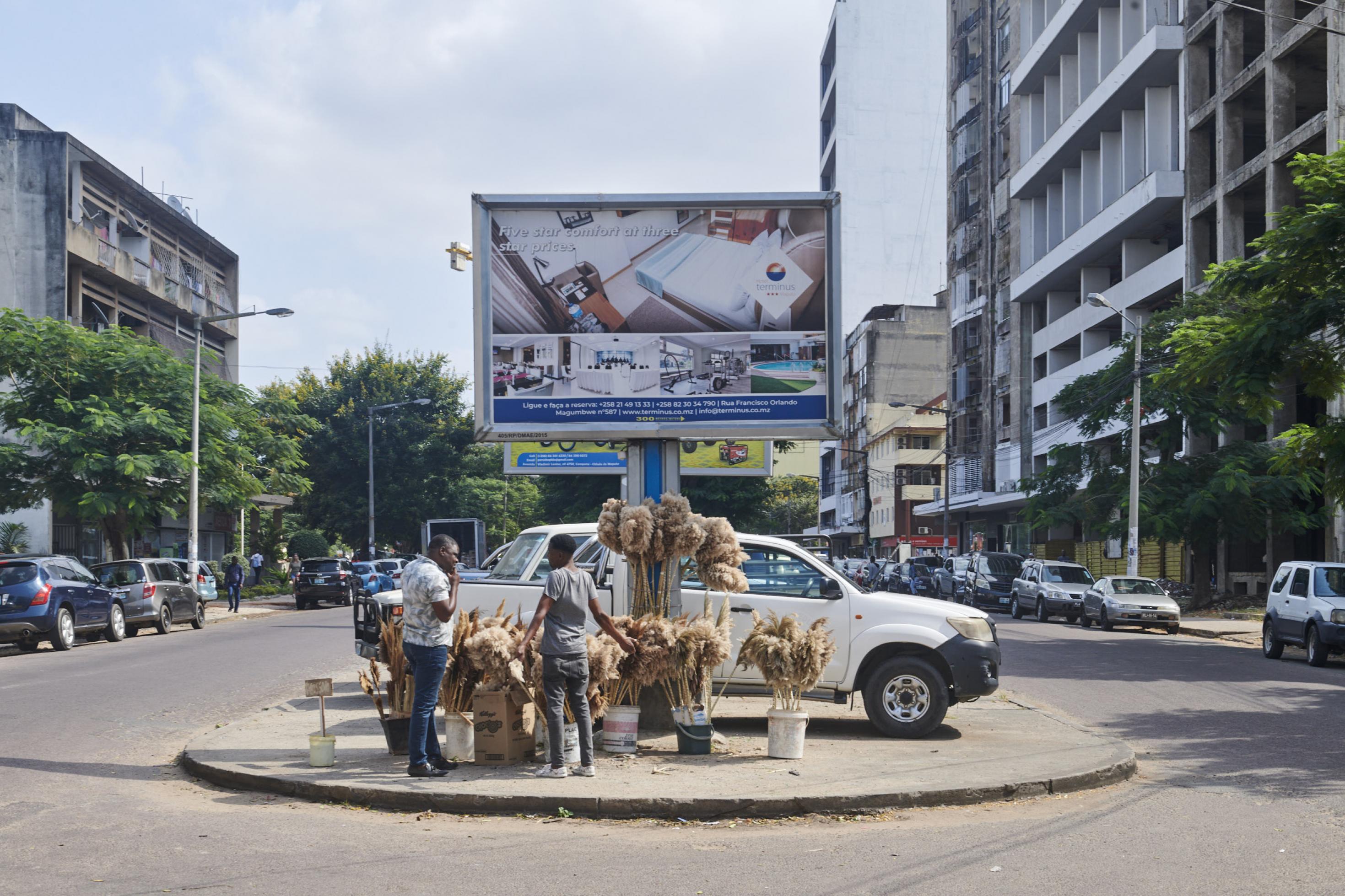 Vor einem großen Werbeplakat auf einer Kreuzung diskutiert ein Straßenhändler mit einem Kunden, er verkauft Staubwedel