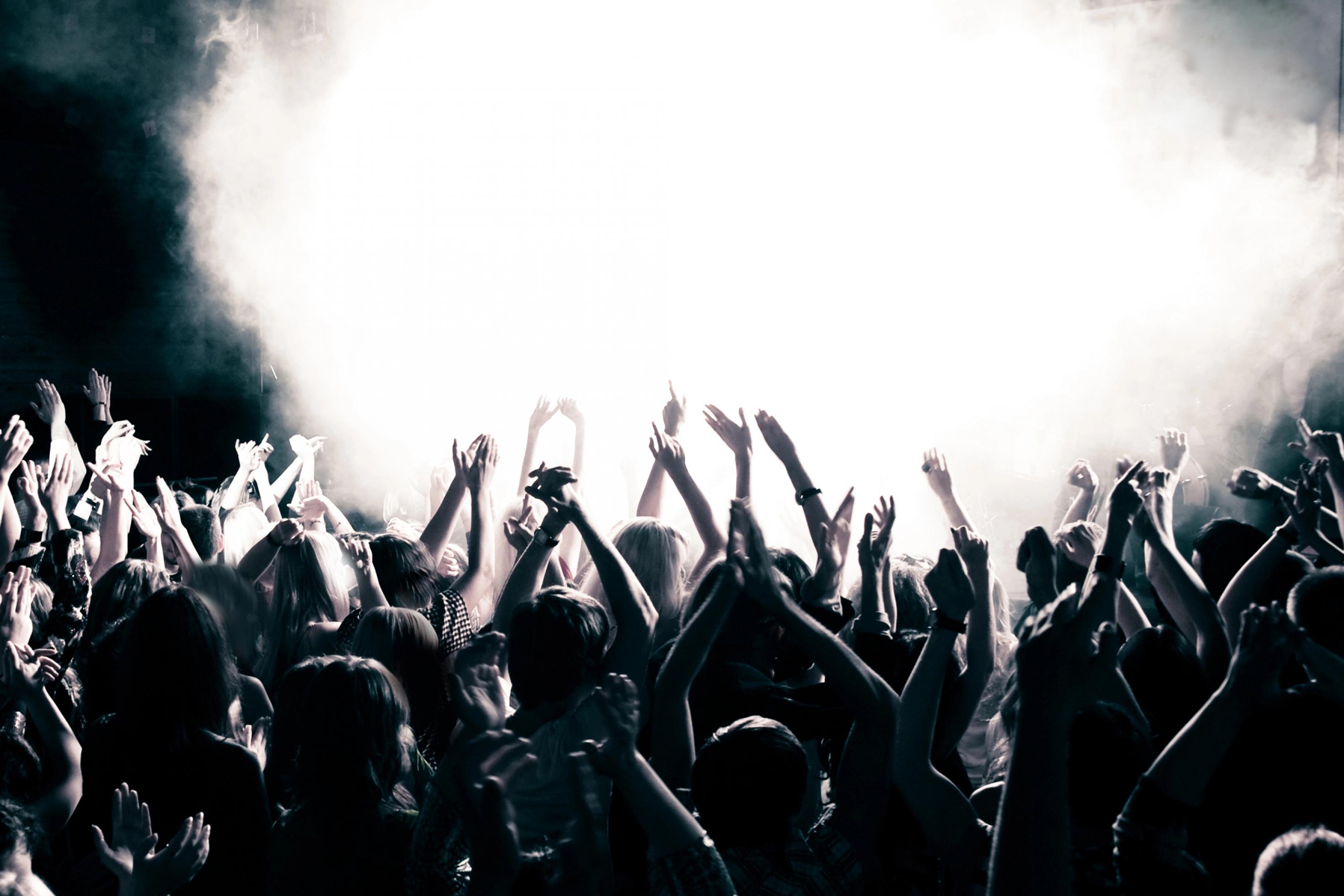 Das Schwarz-Weiß-Foto zeigt die Silhouetten eine Menge junger Menschen, die während einer Musikveranstaltung in einem Saal enthusiastisch tanzen und die Arme hochrecken – im Hintergrund eine gleißend helle, von Scheinwerfern angeleuchtete Nebelblase.