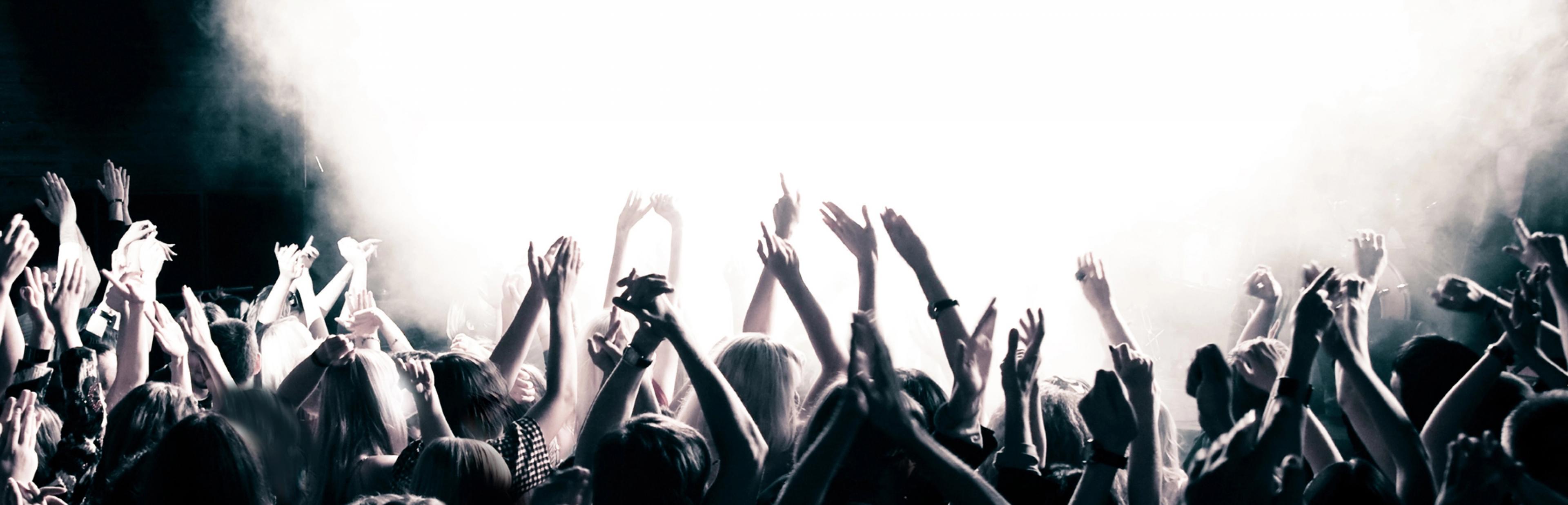 Das Schwarz-Weiß-Foto zeigt die Silhouetten eine Menge junger Menschen, die während einer Musikveranstaltung in einem Saal enthusiastisch tanzen und die Arme hochrecken – im Hintergrund eine gleißend helle, von Scheinwerfern angeleuchtete Nebelblase.