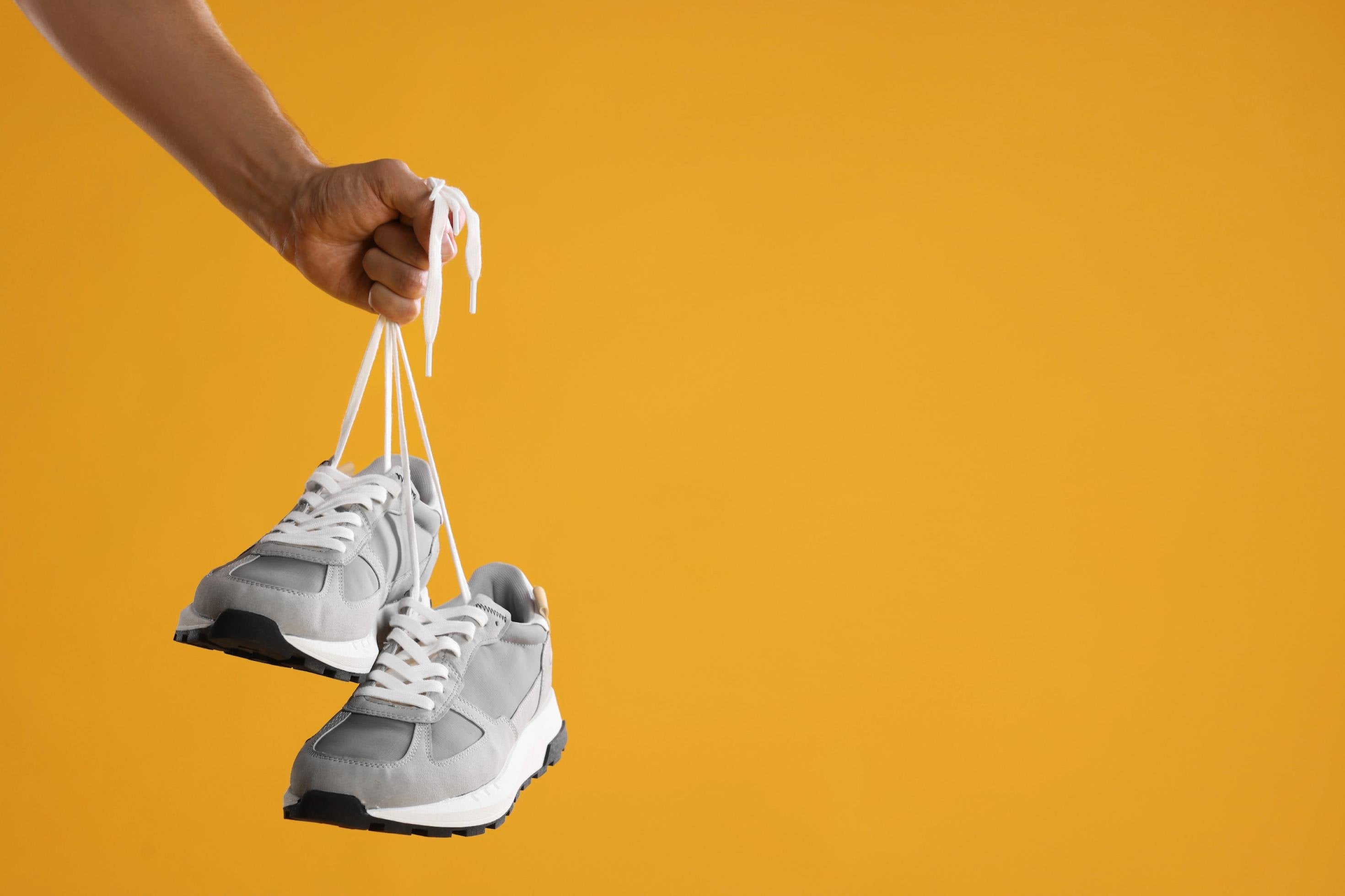 Eine Hand hält helle Sportschuhe vor einer gelben Wand hoch.